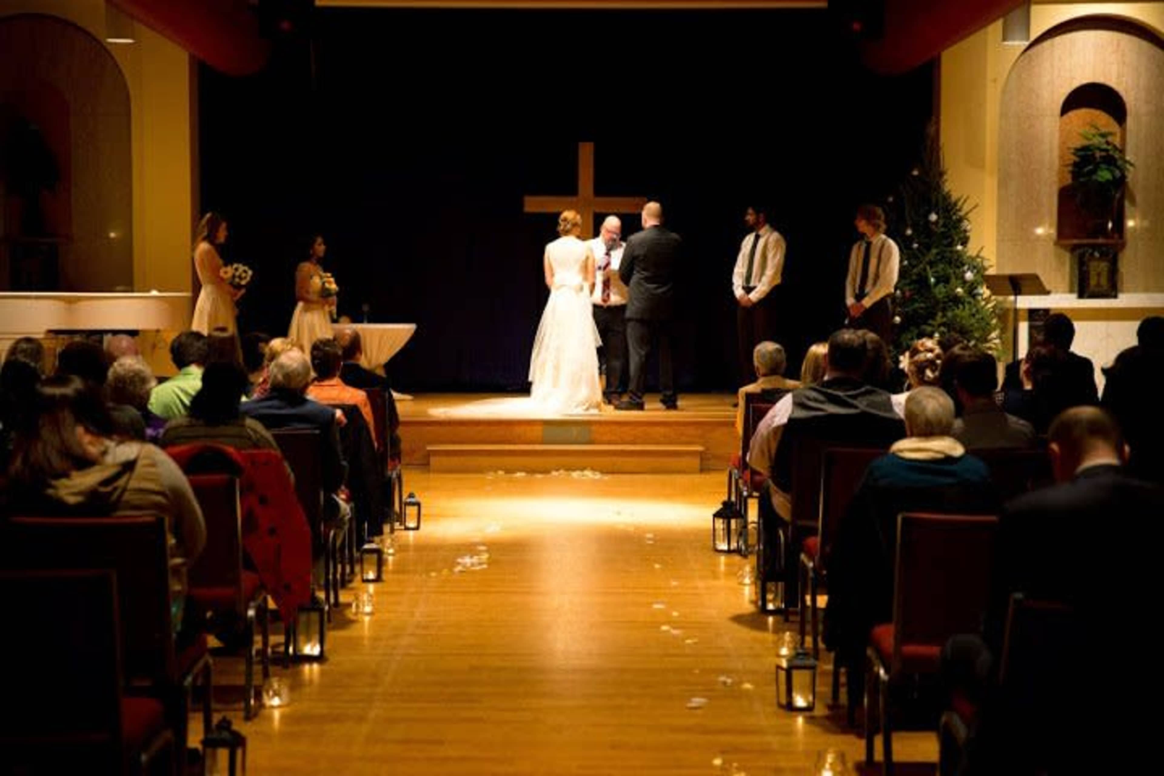 A wedding ceremony is taking place on a stage in a church, with the couple standing before a cross and guests seated in rows facing them.