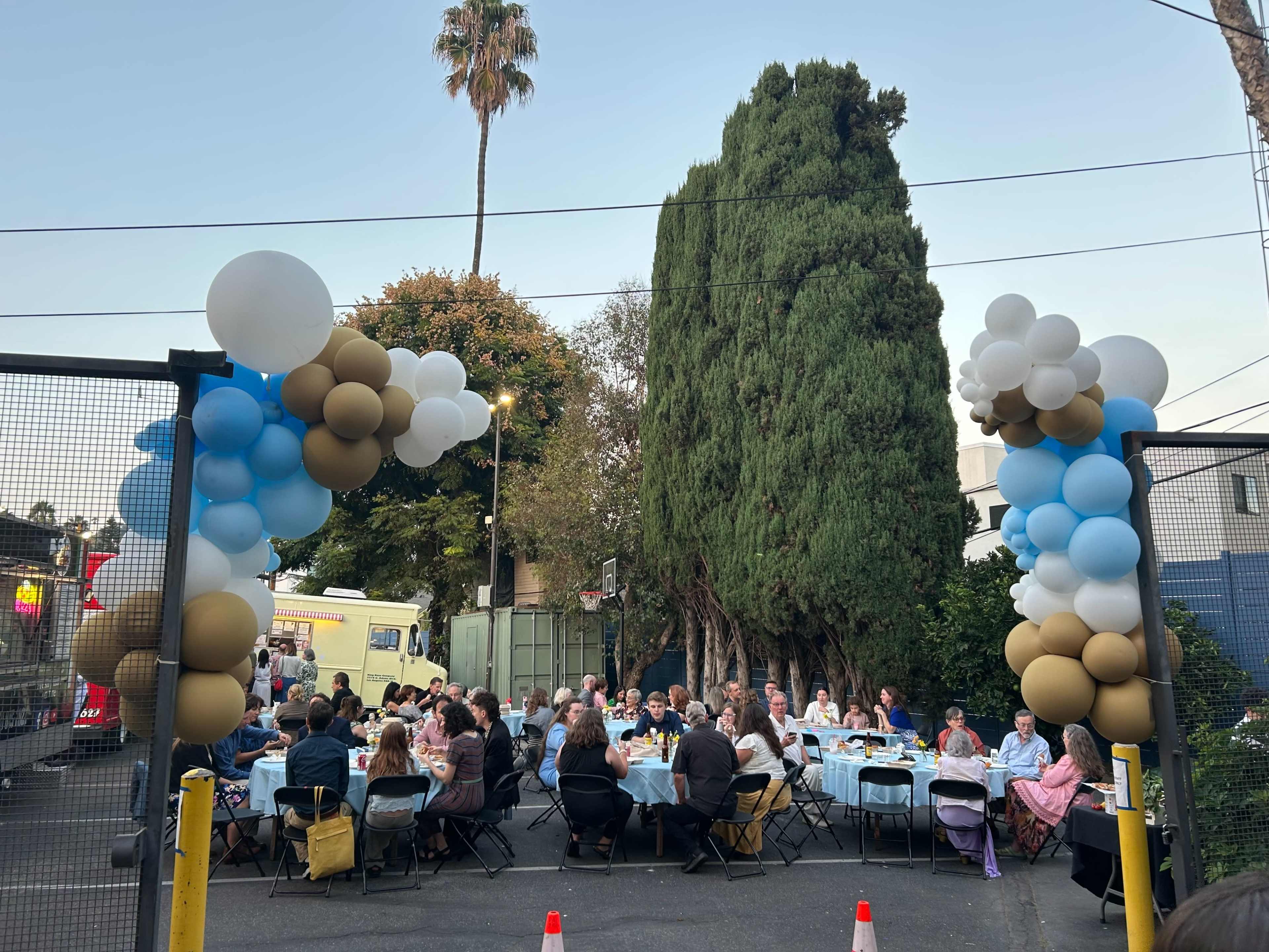 A large outdoor gathering is set up with tables and chairs arranged under trees, decorated with balloon arches, as people enjoy a meal together.