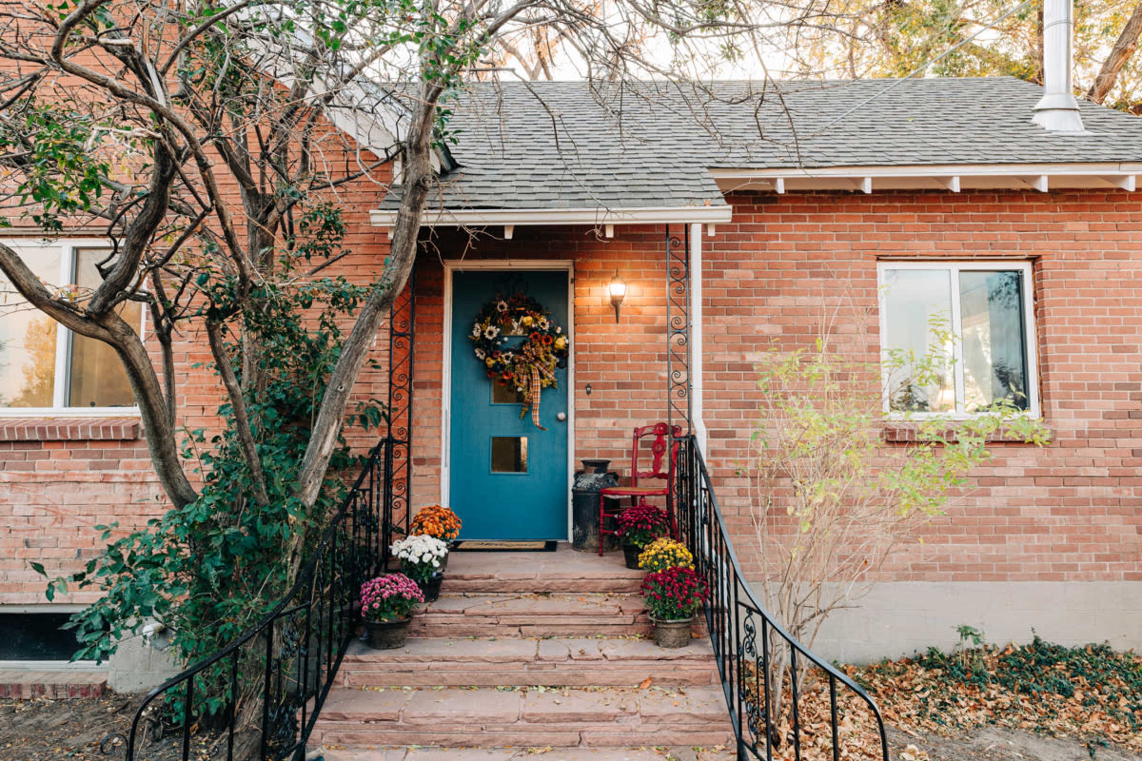 The front entrance of a brick house features a blue door adorned with a wreath, flanked by potted flowers on stone steps.