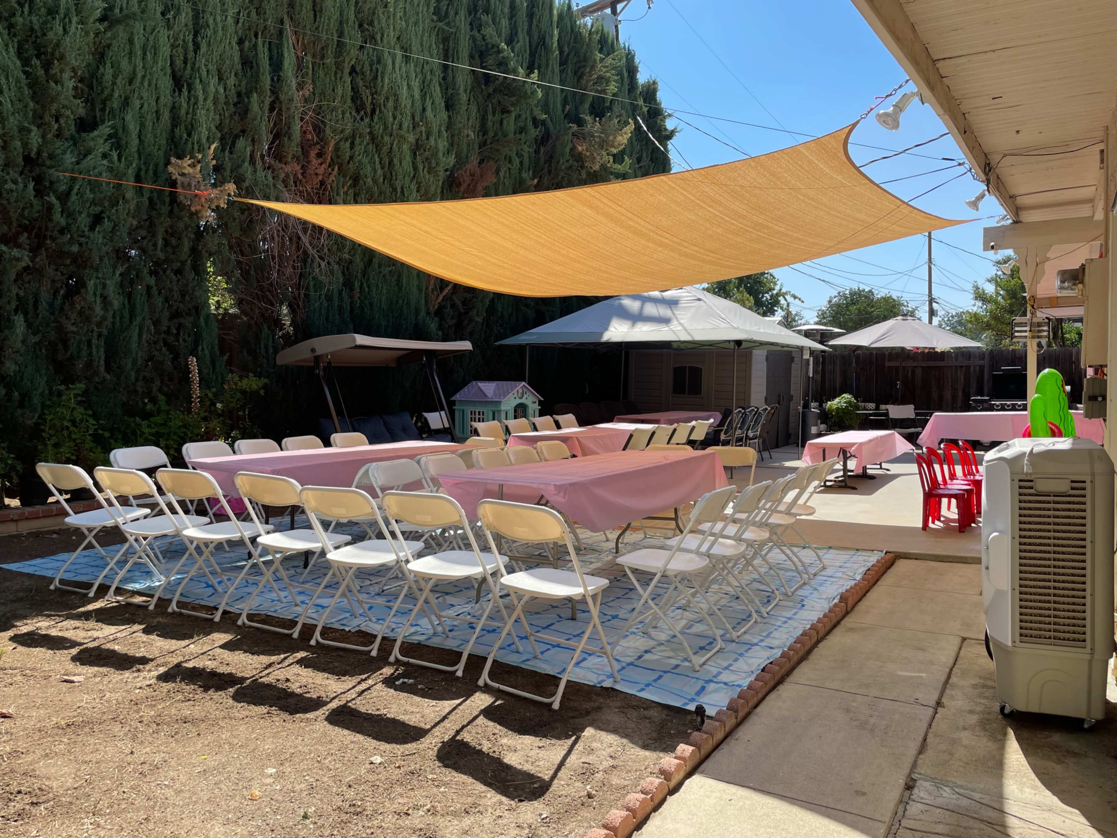 A large outdoor space is set up with several long tables covered in pink tablecloths, surrounded by white chairs, and shaded by a large fabric canopy.