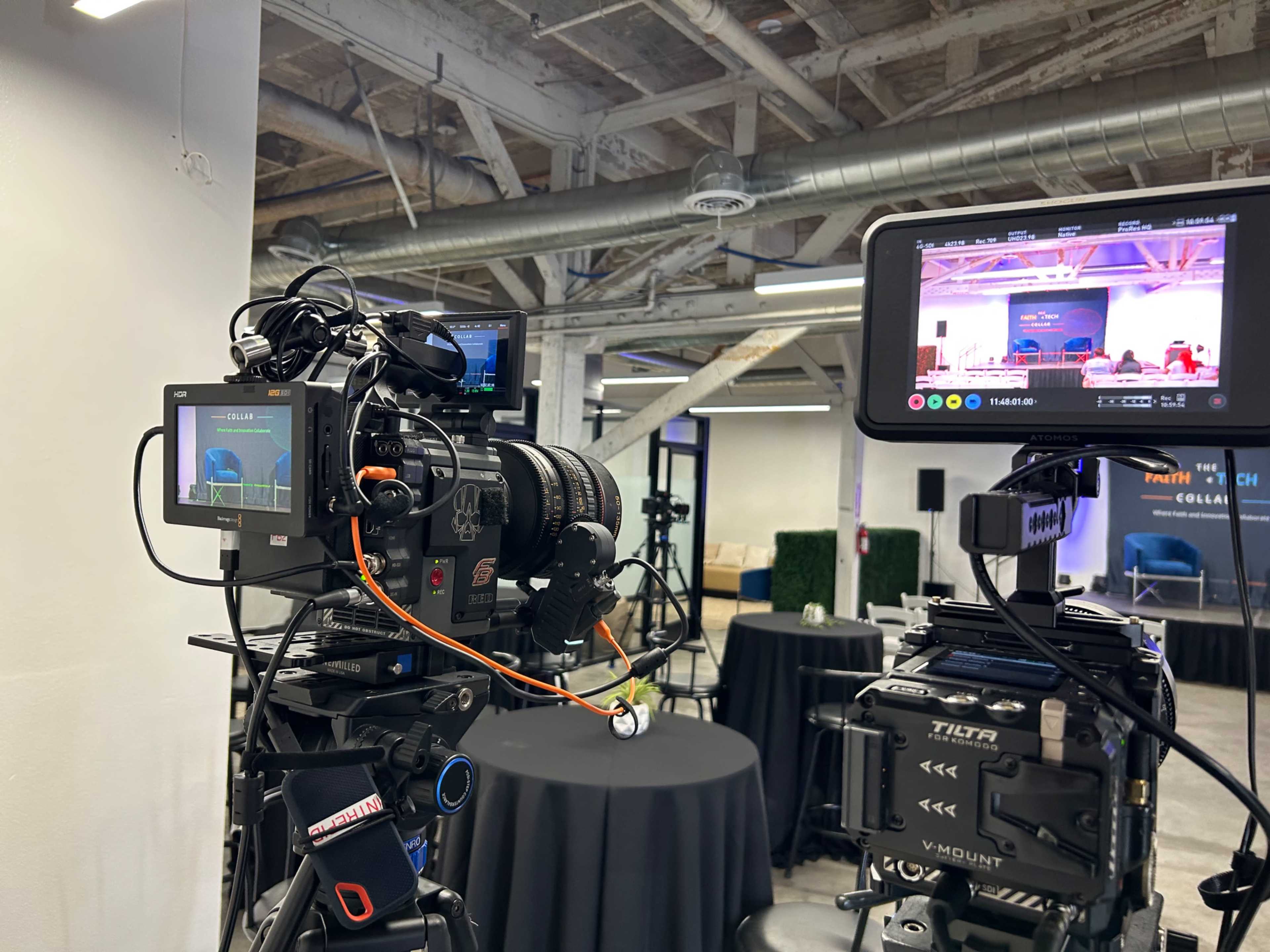 The image shows two professional cameras set up on tripods in a spacious indoor venue with a few tables and equipment in the background.