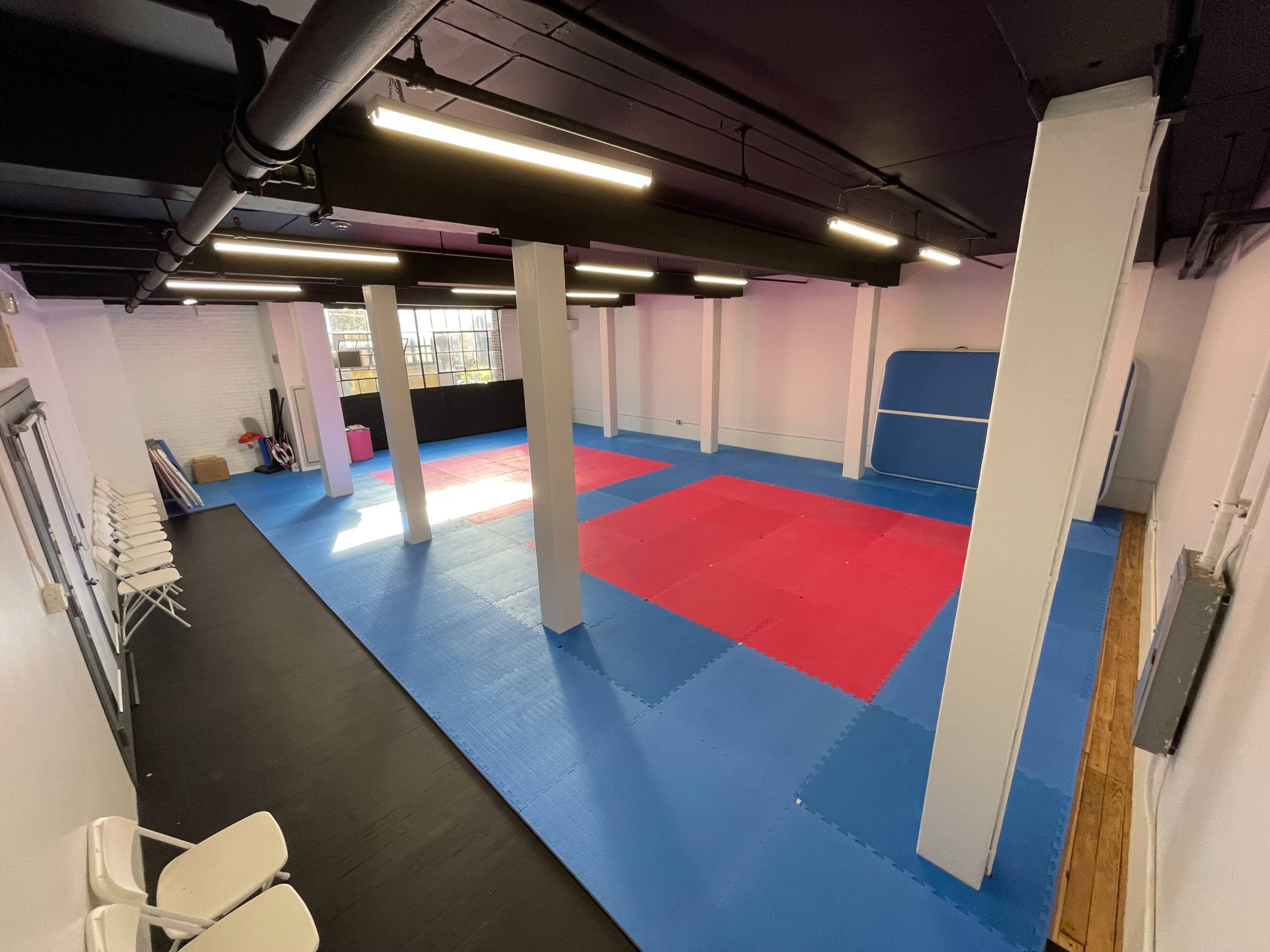 A spacious martial arts training room with red and blue mats, white columns, and overhead lighting.