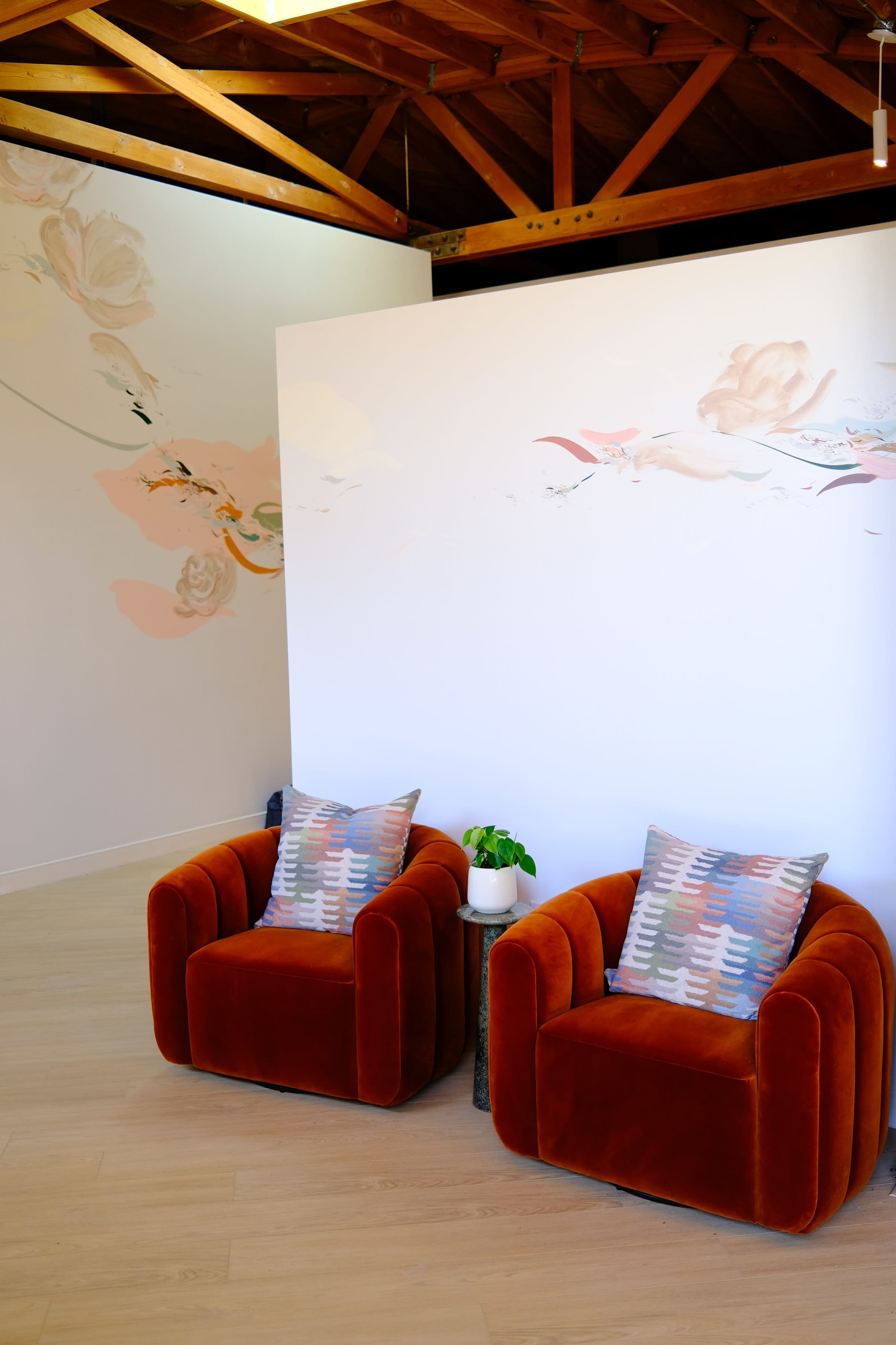 Two orange velvet chairs with patterned cushions are positioned in front of a white wall adorned with floral artwork, against a backdrop of exposed wooden beams.