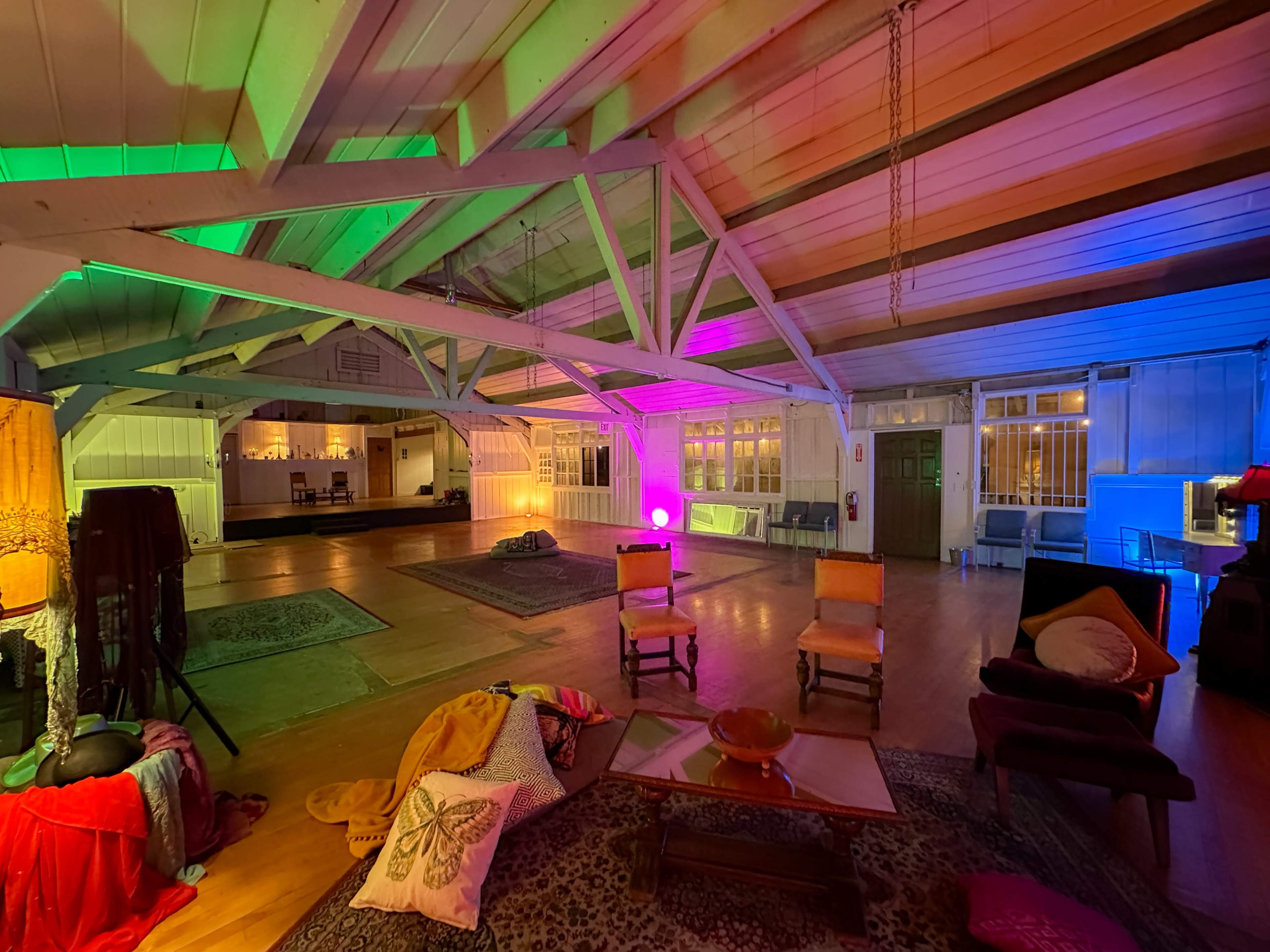 The image shows a large, brightly lit room with colorful lighting, wooden beams, and various seating arrangements scattered across the floor.