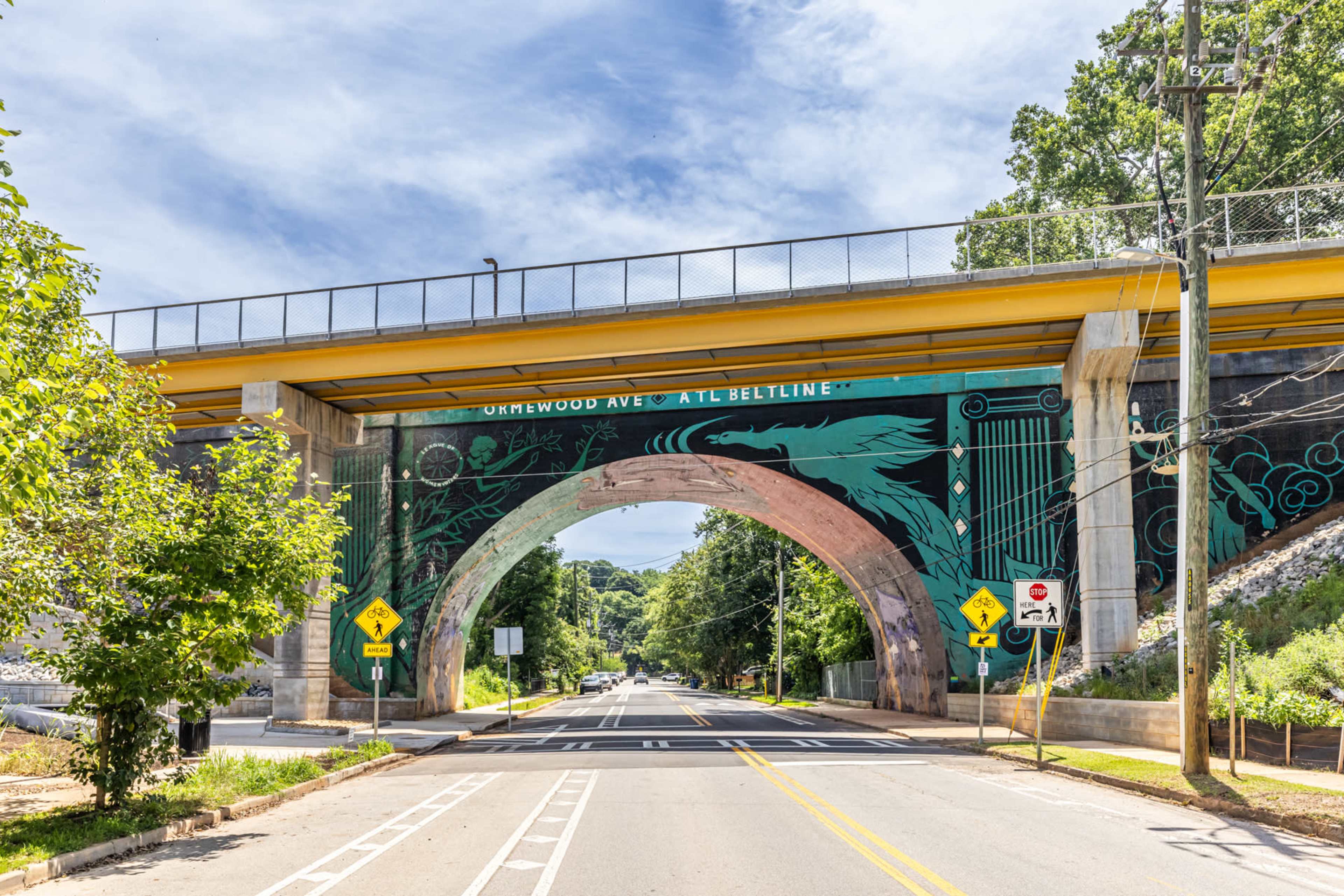 A colorful mural decorates the underside of a bridge that spans a street lined with trees.