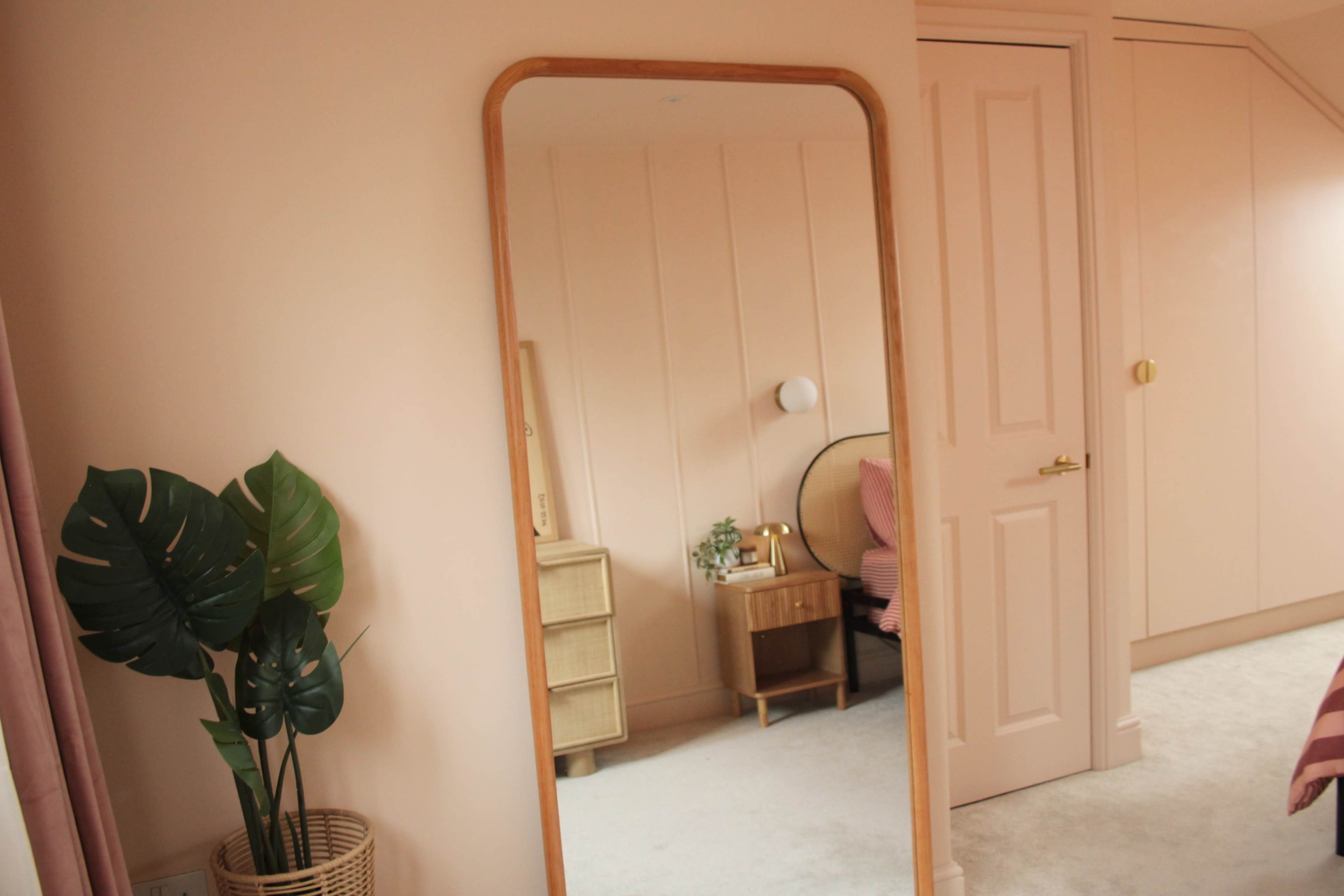 A large, framed mirror reflects a softly lit bedroom with a plant, a small wooden nightstand, and a closet.