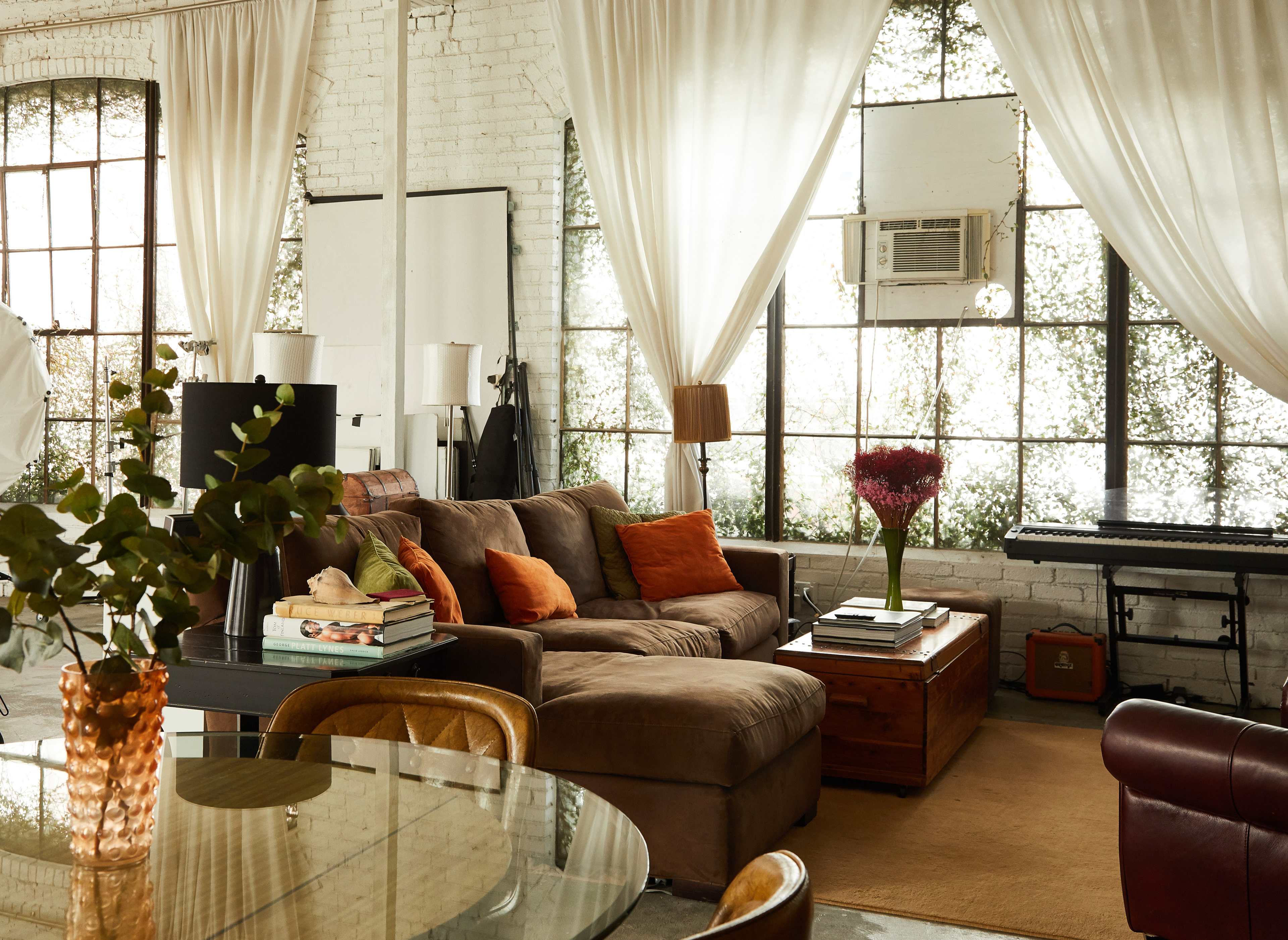 The image shows a stylish living area with a brown sofa, decorative pillows, a coffee table, and large windows covered with sheer curtains.