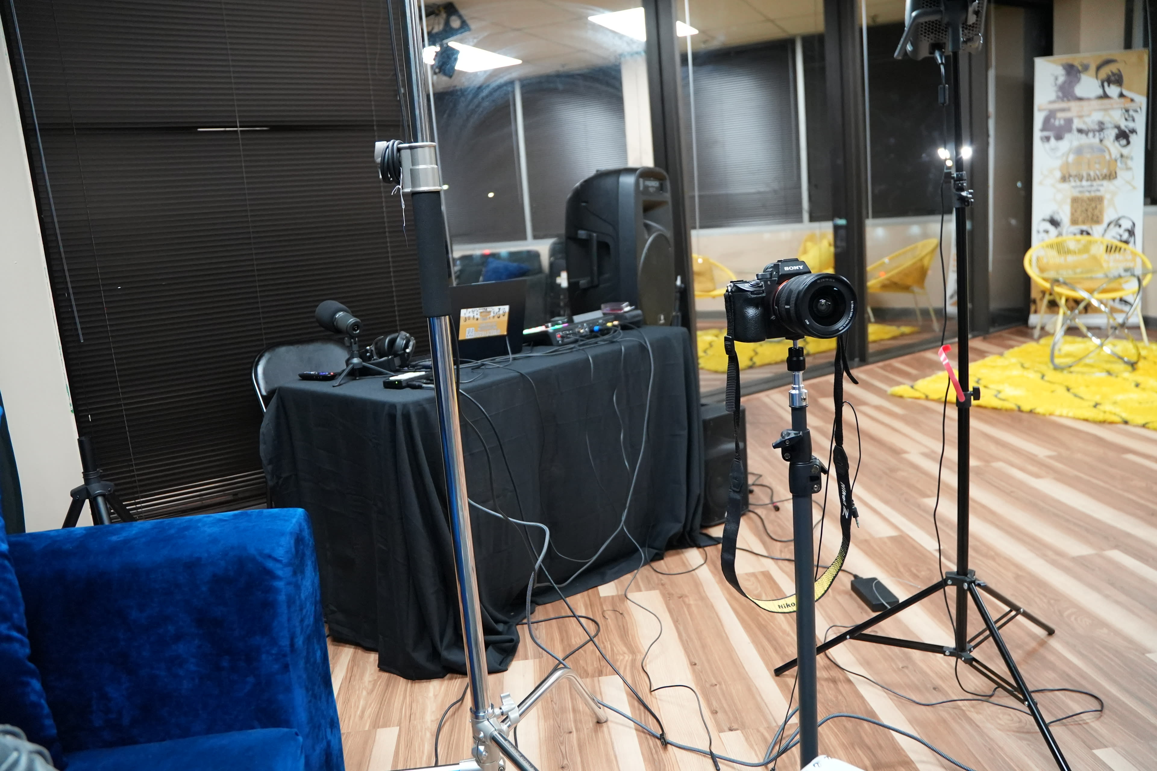 The image shows a room set up for recording, featuring a camera on a tripod, a microphone, and a black table with audio equipment.