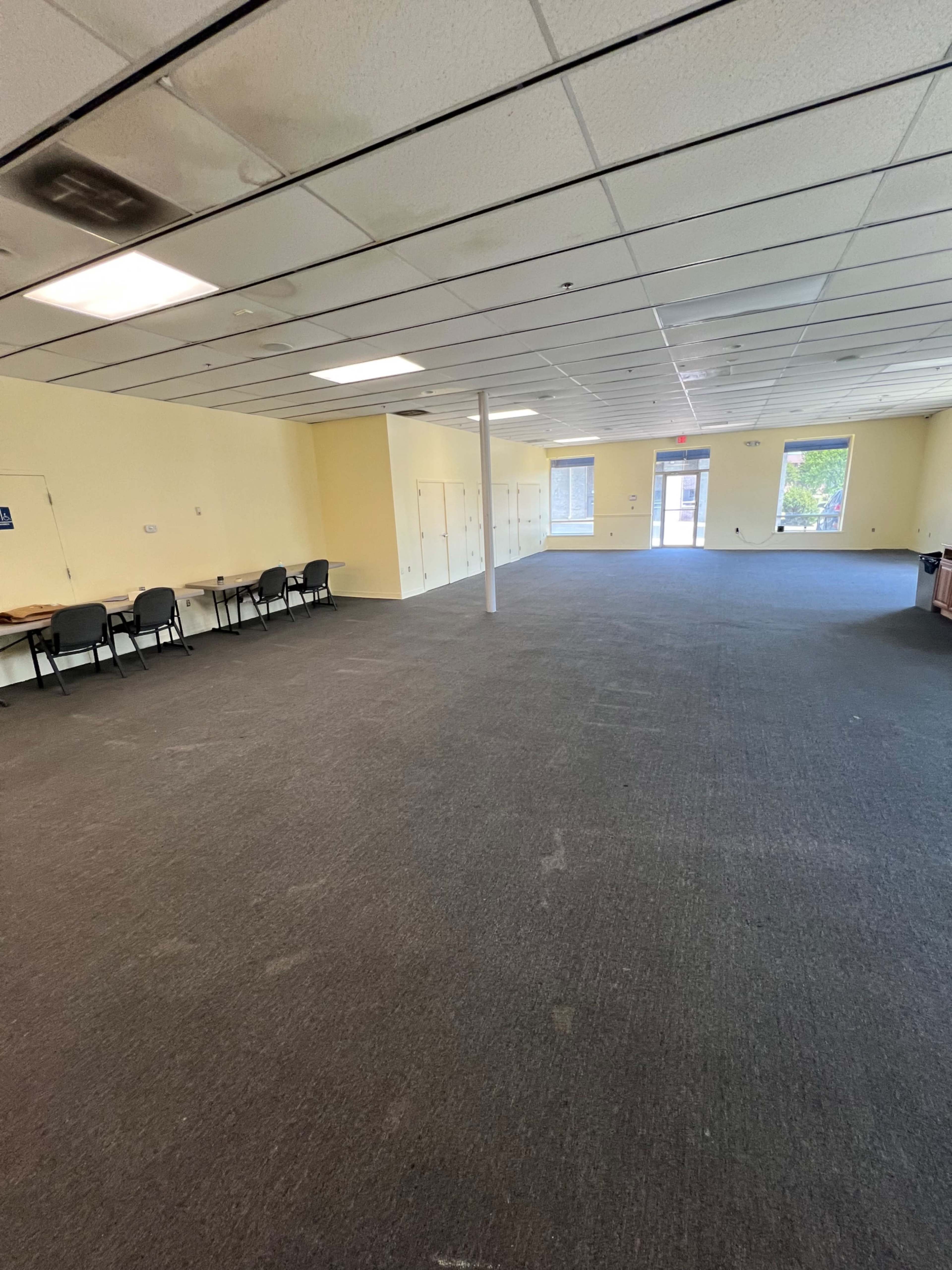 The image shows a spacious, empty room with yellow walls, several tables and chairs along one side, and a door leading to an outdoor area.