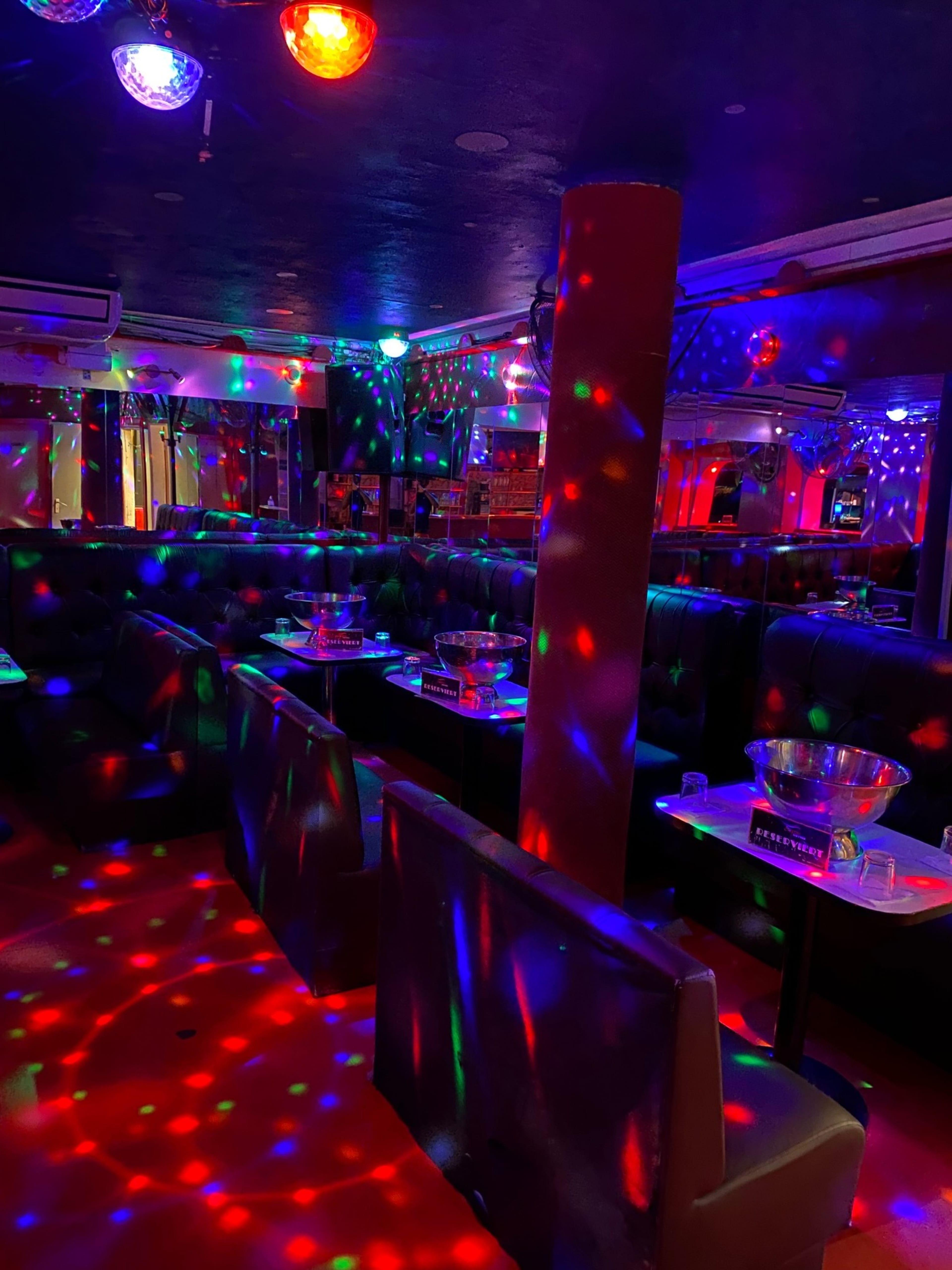 A dimly lit lounge features dark seating, multicolored disco lights, and mirrored walls.