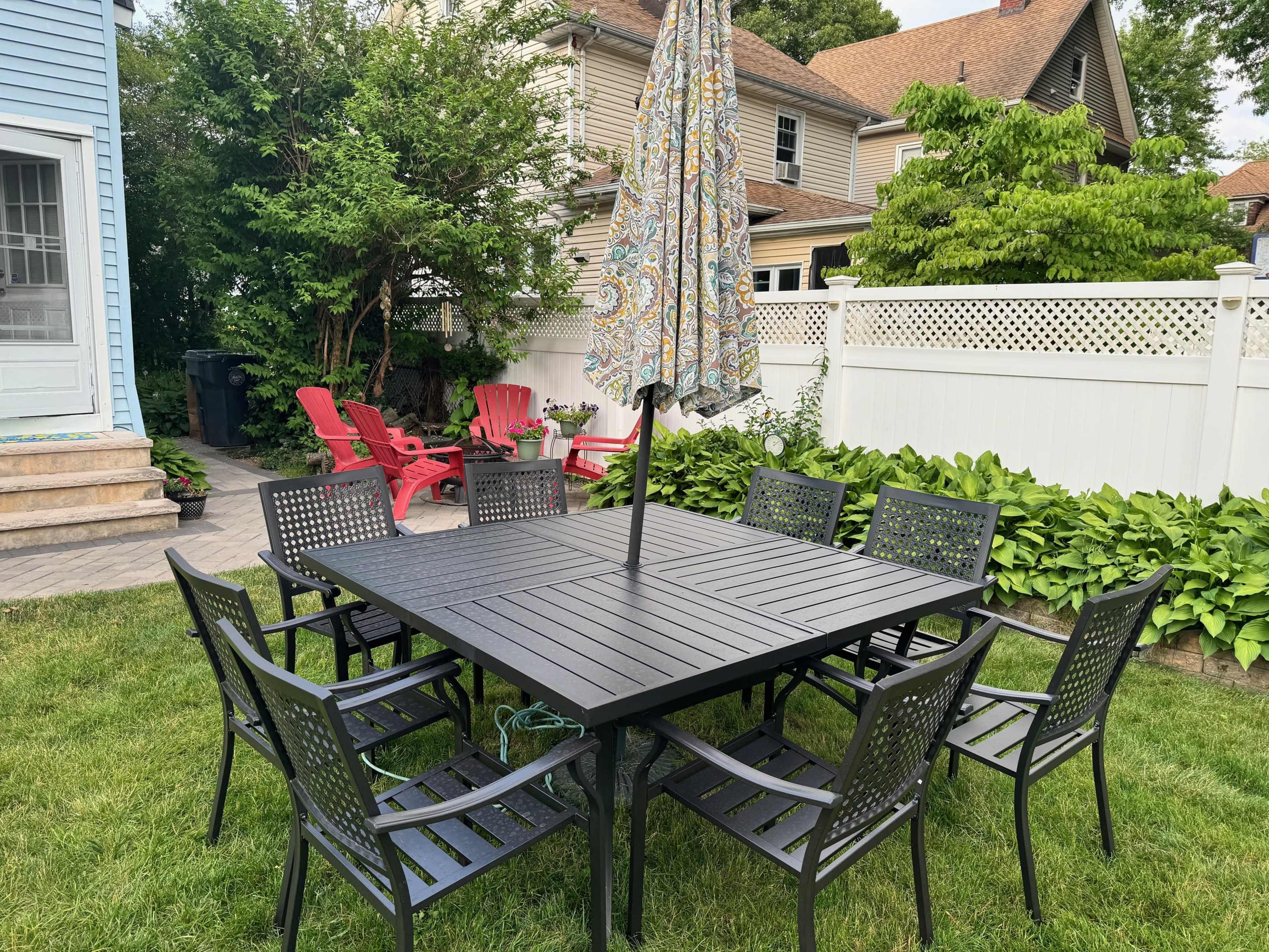 A square patio table with eight black chairs and an umbrella is situated on a grassy area surrounded by colorful plants and a white fence.
