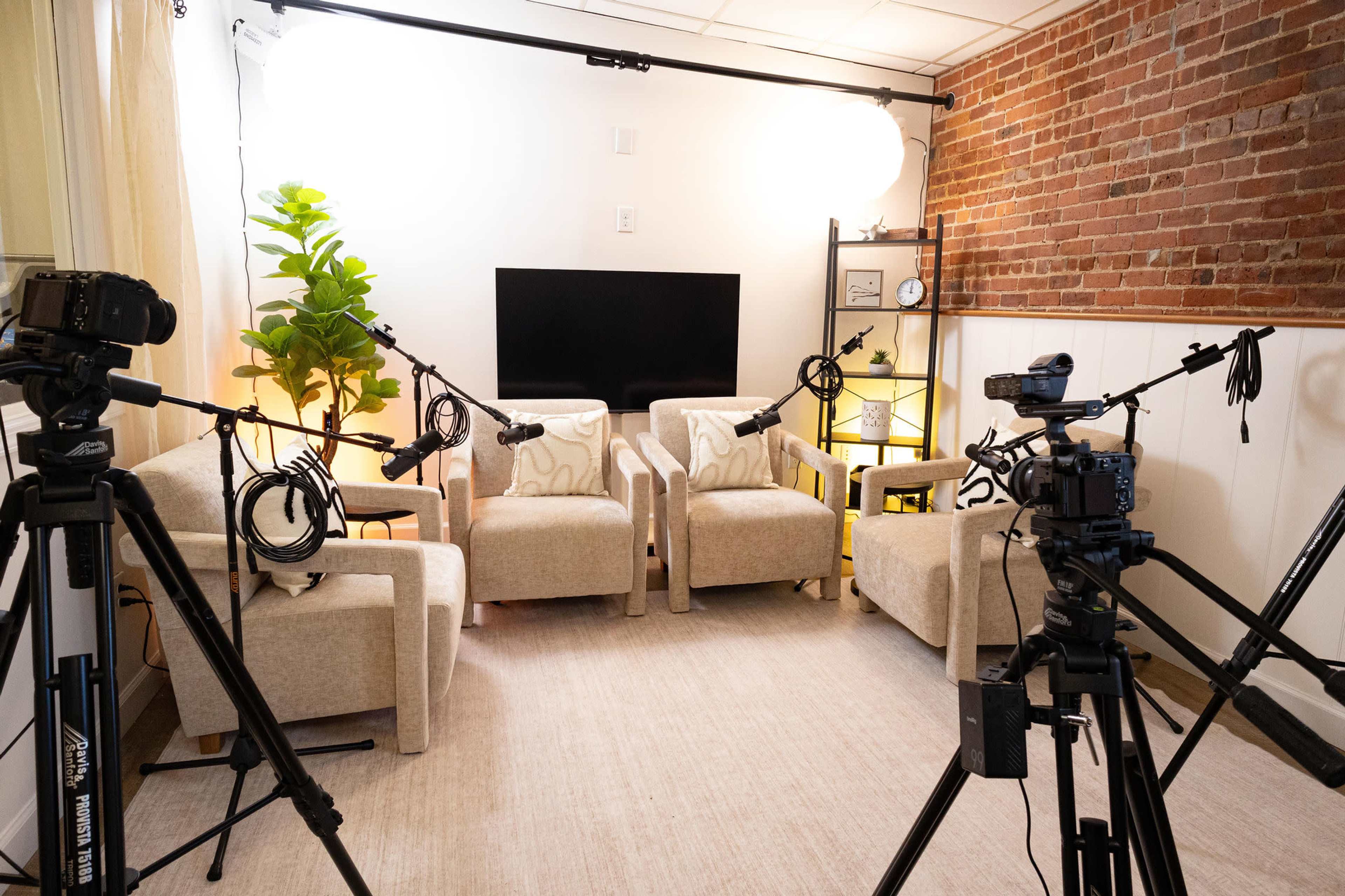 The image shows a professionally set up interview space featuring four armchairs, a television screen on the wall, and multiple cameras and microphones positioned around the area.