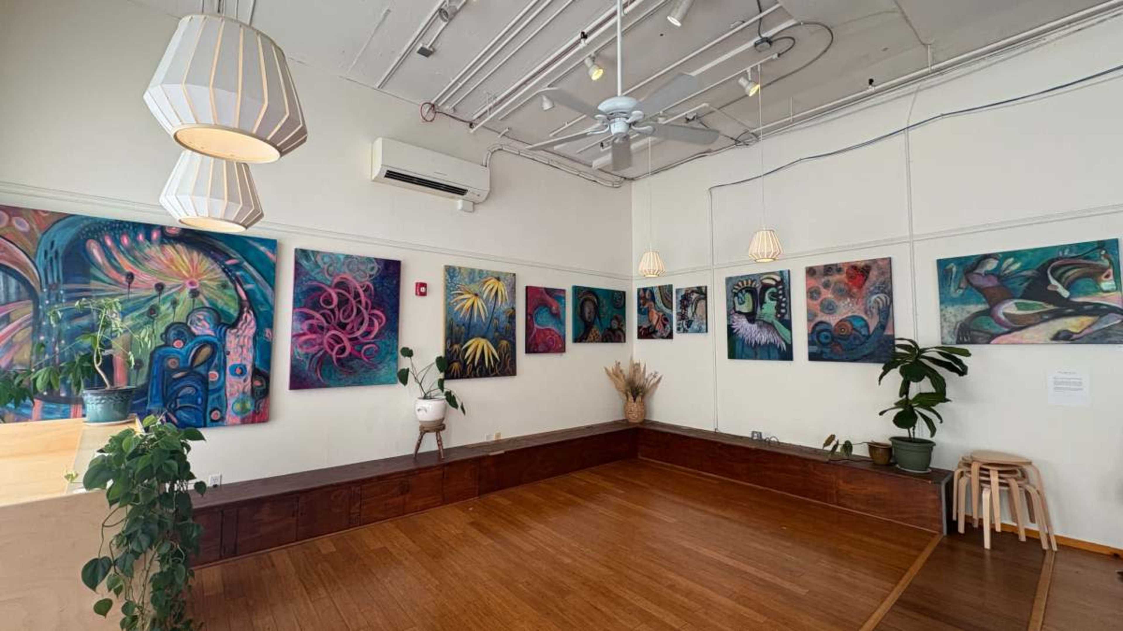 The image shows an art gallery with colorful paintings displayed on the walls, wooden flooring, and decorative plants.