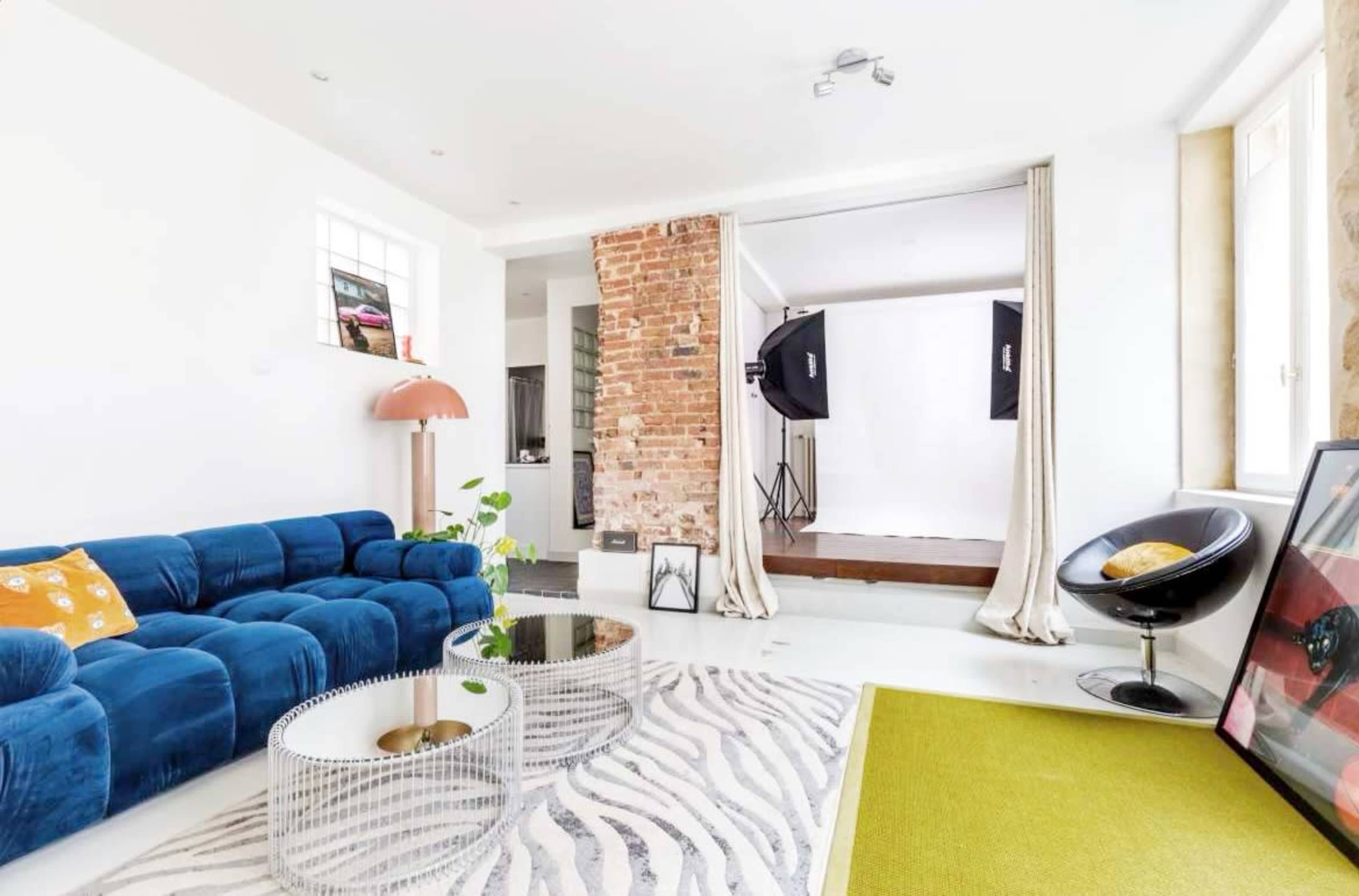 The image shows a bright living room featuring a blue modular sofa, a modern coffee table, and a photography setup with softbox lights in the background.