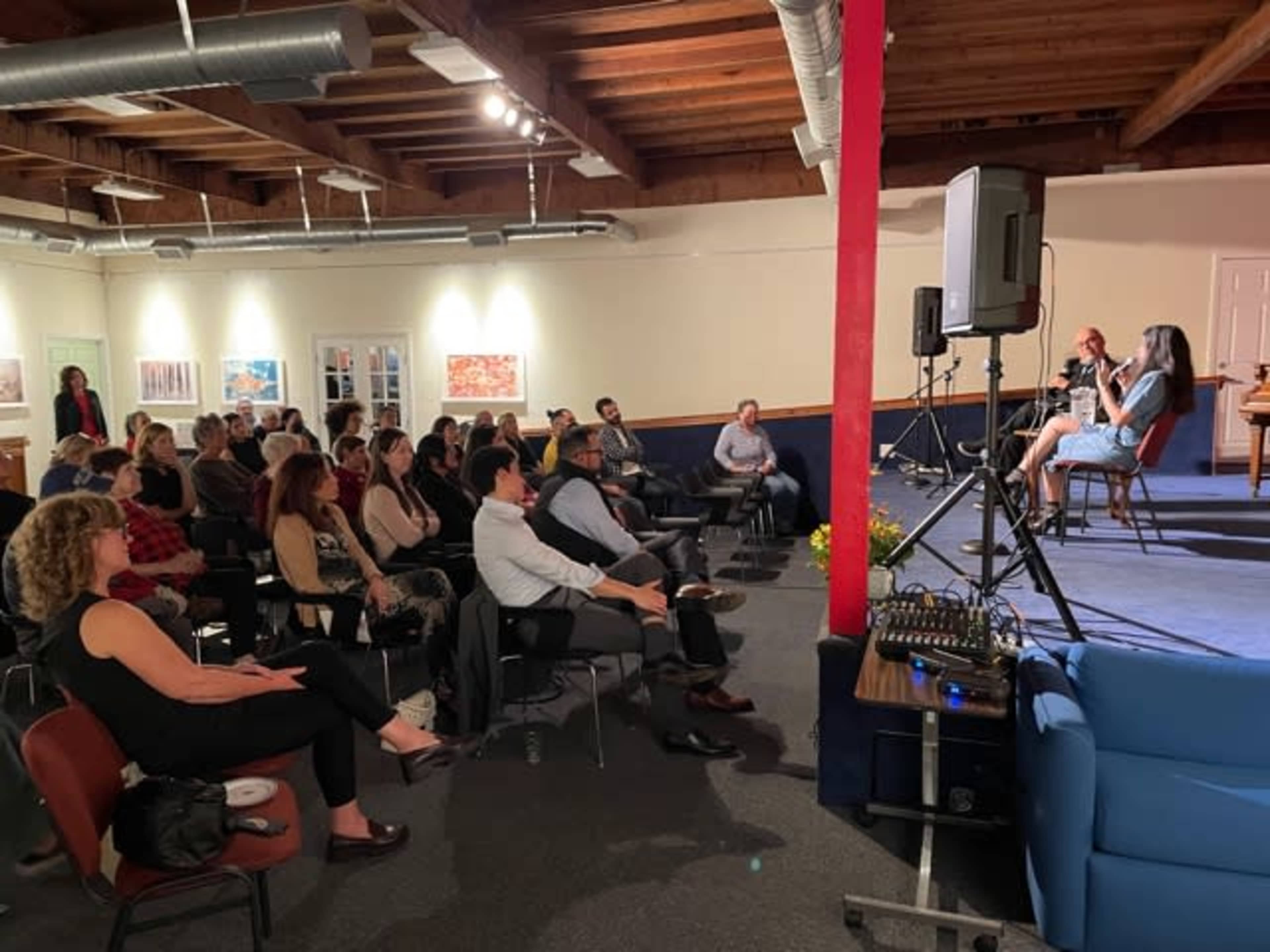 A crowd is seated in a room facing a stage where two speakers are engaged in a discussion, while a sound system is set up nearby.
