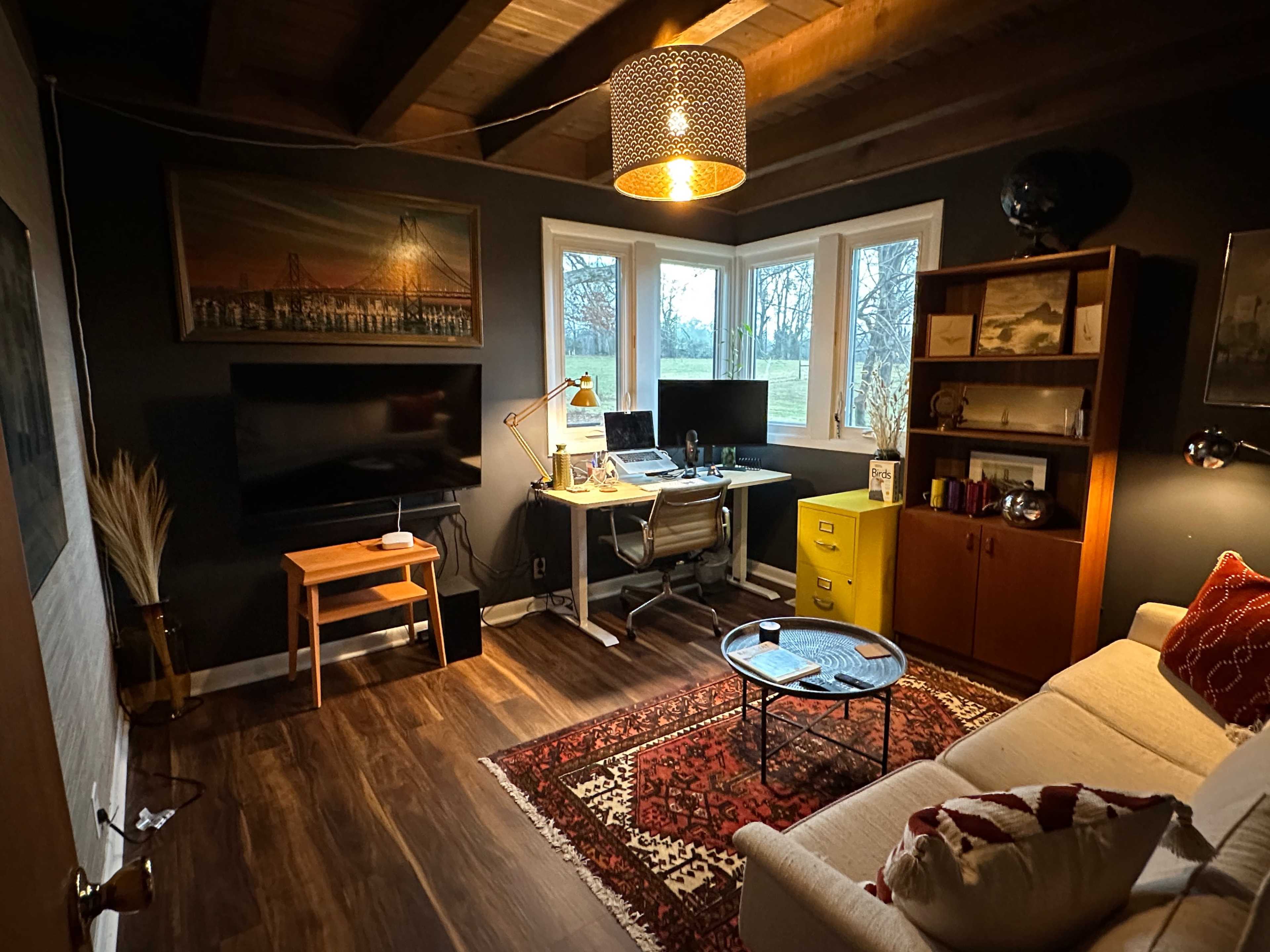 A home office with a desk, two computer screens, a television, a bookshelf, and a sofa, all set against dark walls and wooden beams.