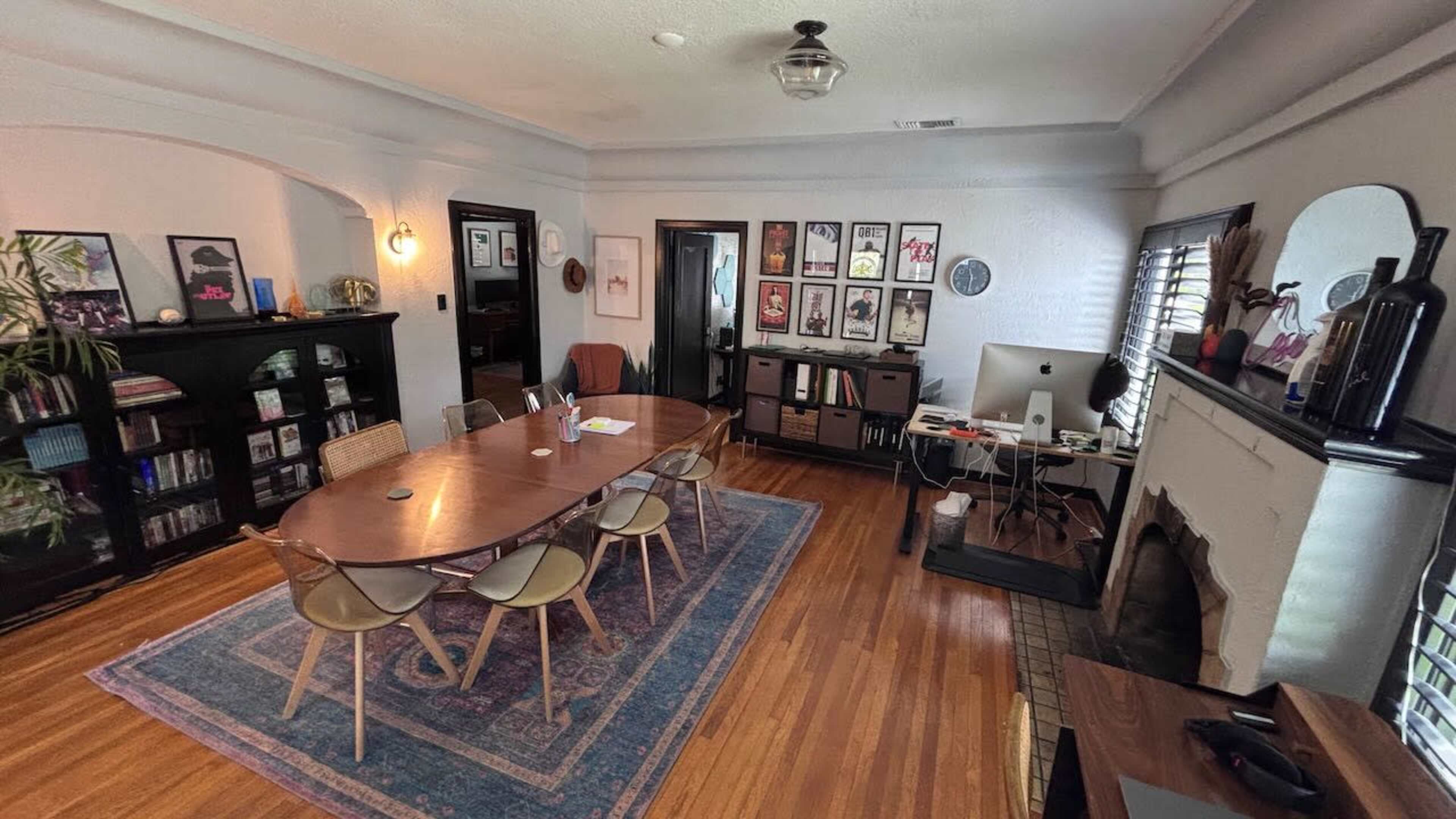 The image shows a modern office space featuring a large wooden table surrounded by chairs, a collection of framed artwork on the walls, and a desk with a computer and office supplies.
