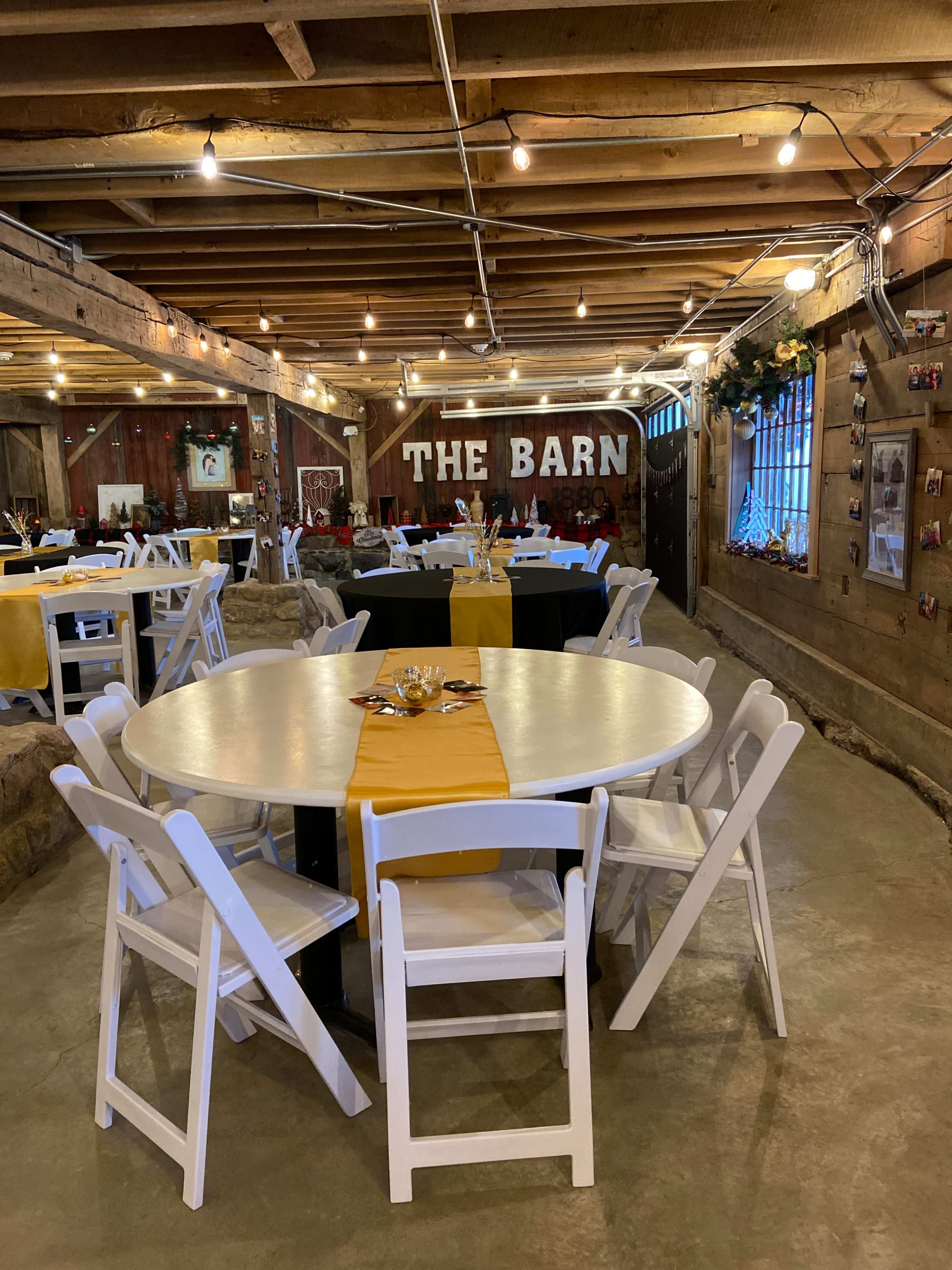 The interior of a rustic barn is set up with round tables covered in white and yellow tablecloths, illuminated by hanging string lights.