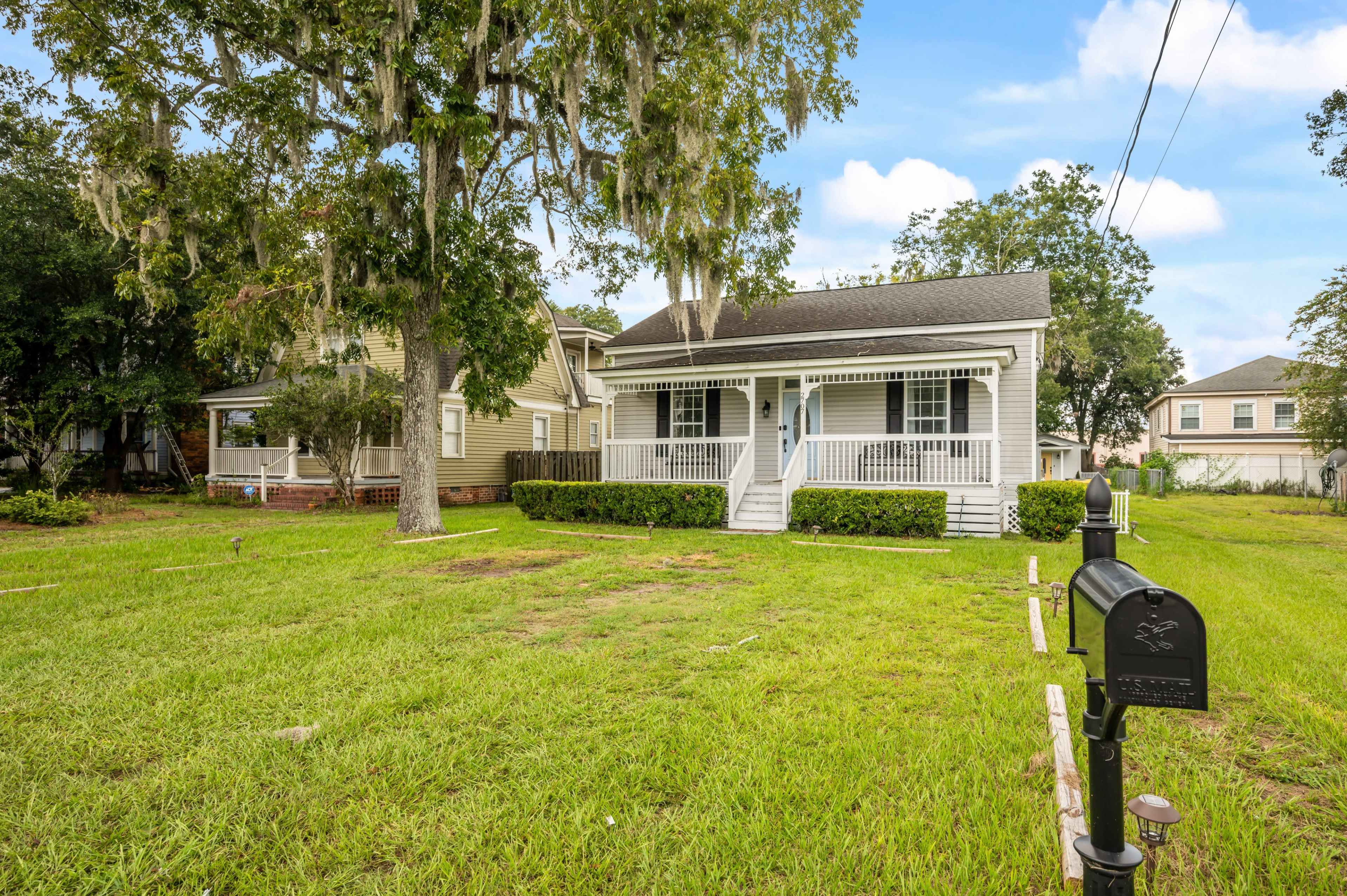 The image shows a white wooden house with a front porch, surrounded by grassy lawns and trees, with a mailbox at the edge of the property.