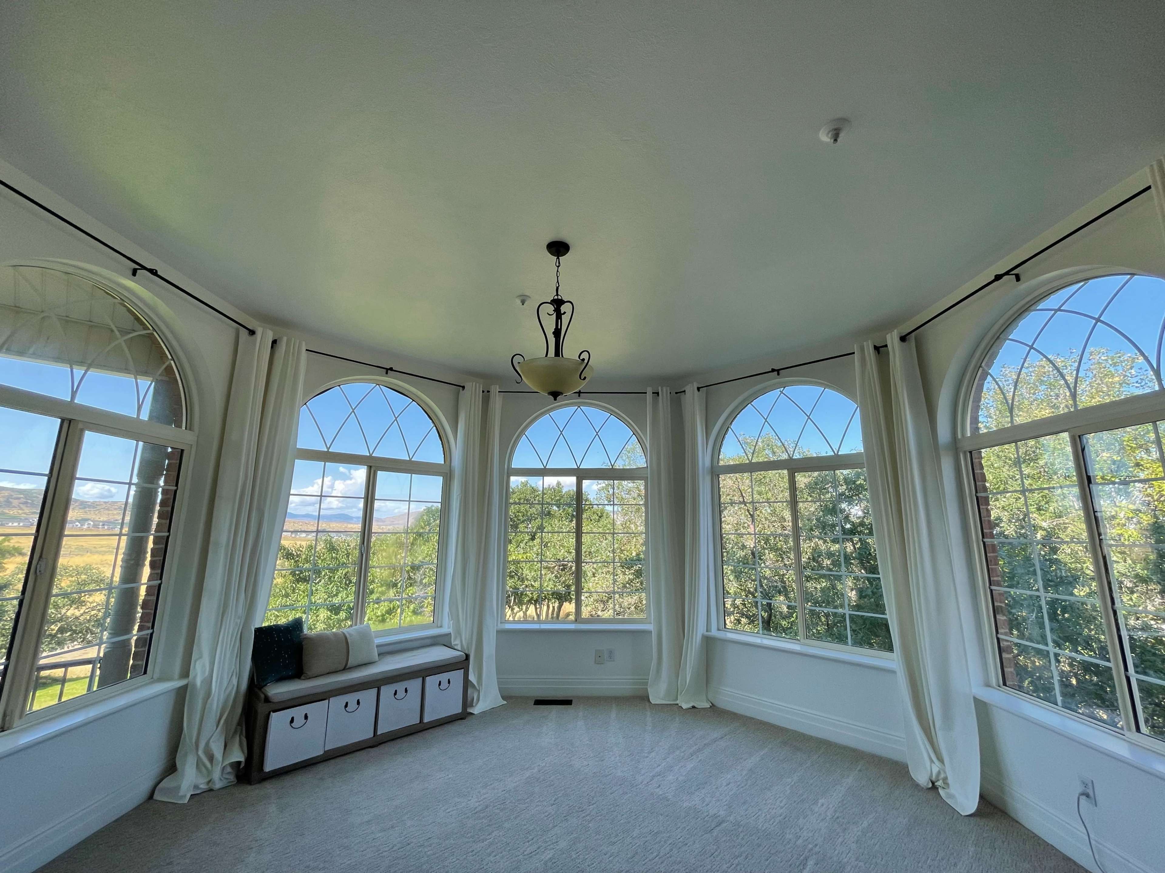 A sunlit room with large arched windows, a light fixture hanging from the ceiling, and a bench with storage underneath, surrounded by greenery outside.