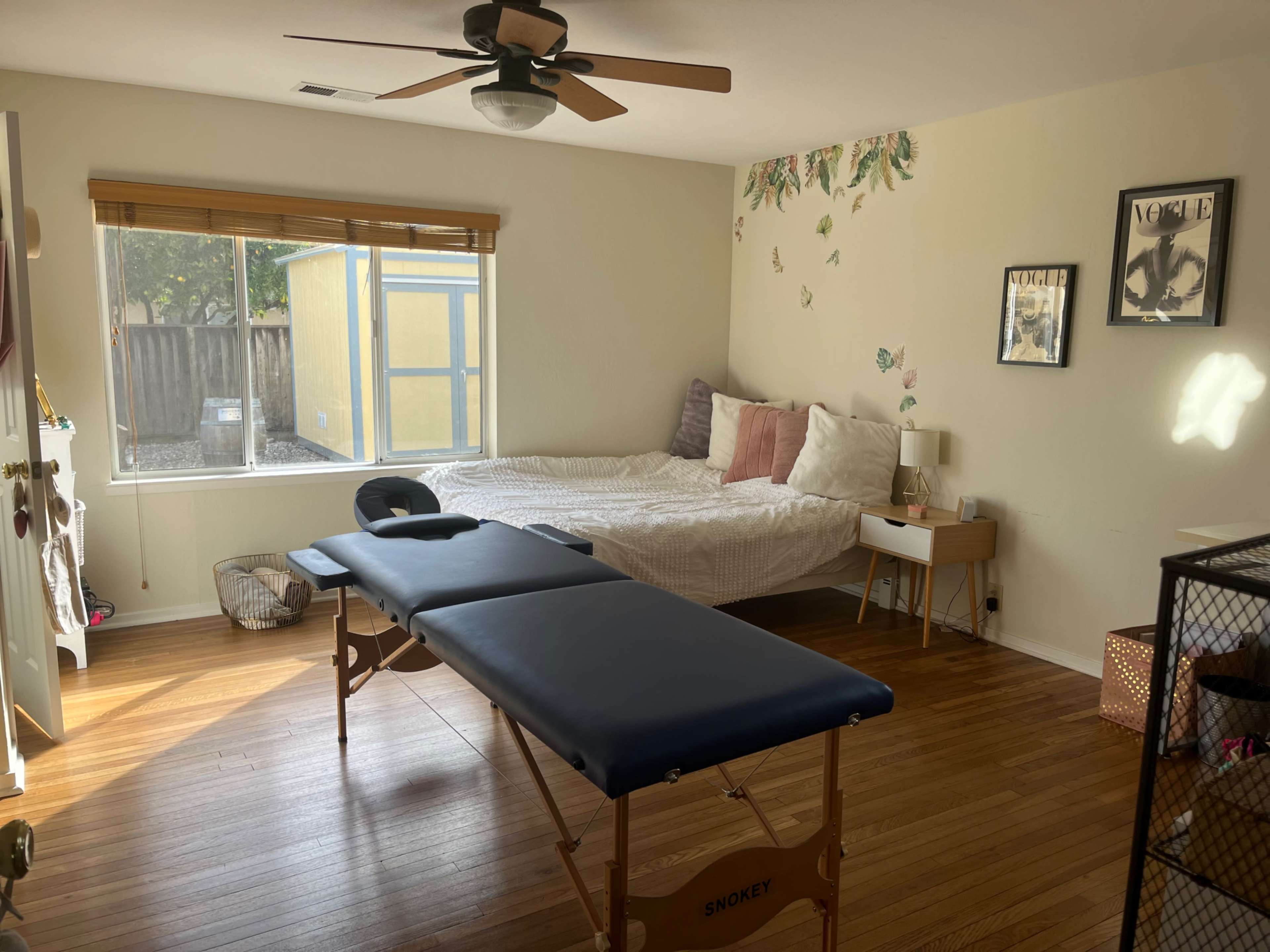 The image shows a bedroom with a bed, a massage table, and a window letting in natural light, decorated with wall art.