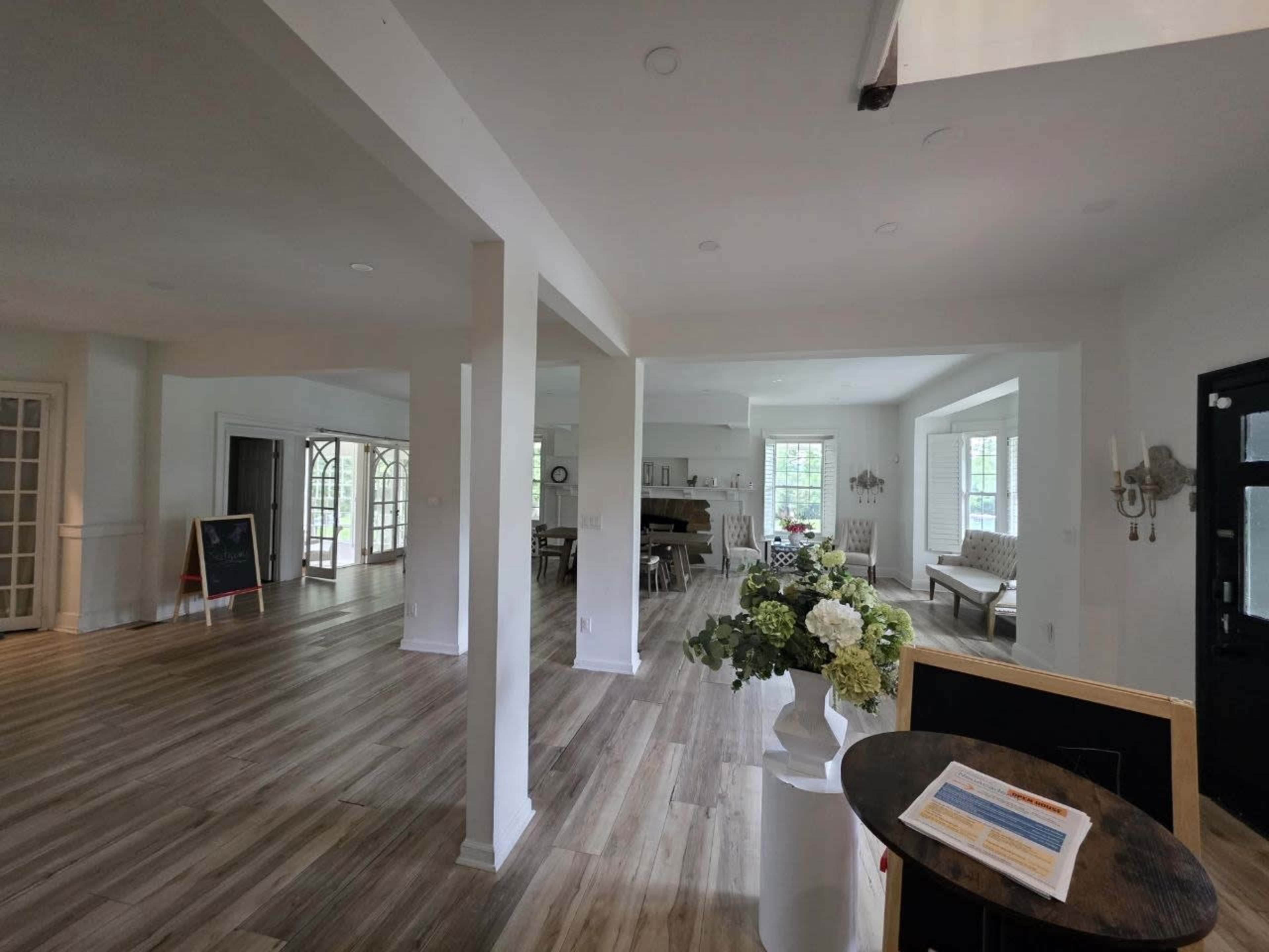 The image shows a spacious, well-lit interior with open areas, wooden flooring, and several furnishings including a floral arrangement and seating.