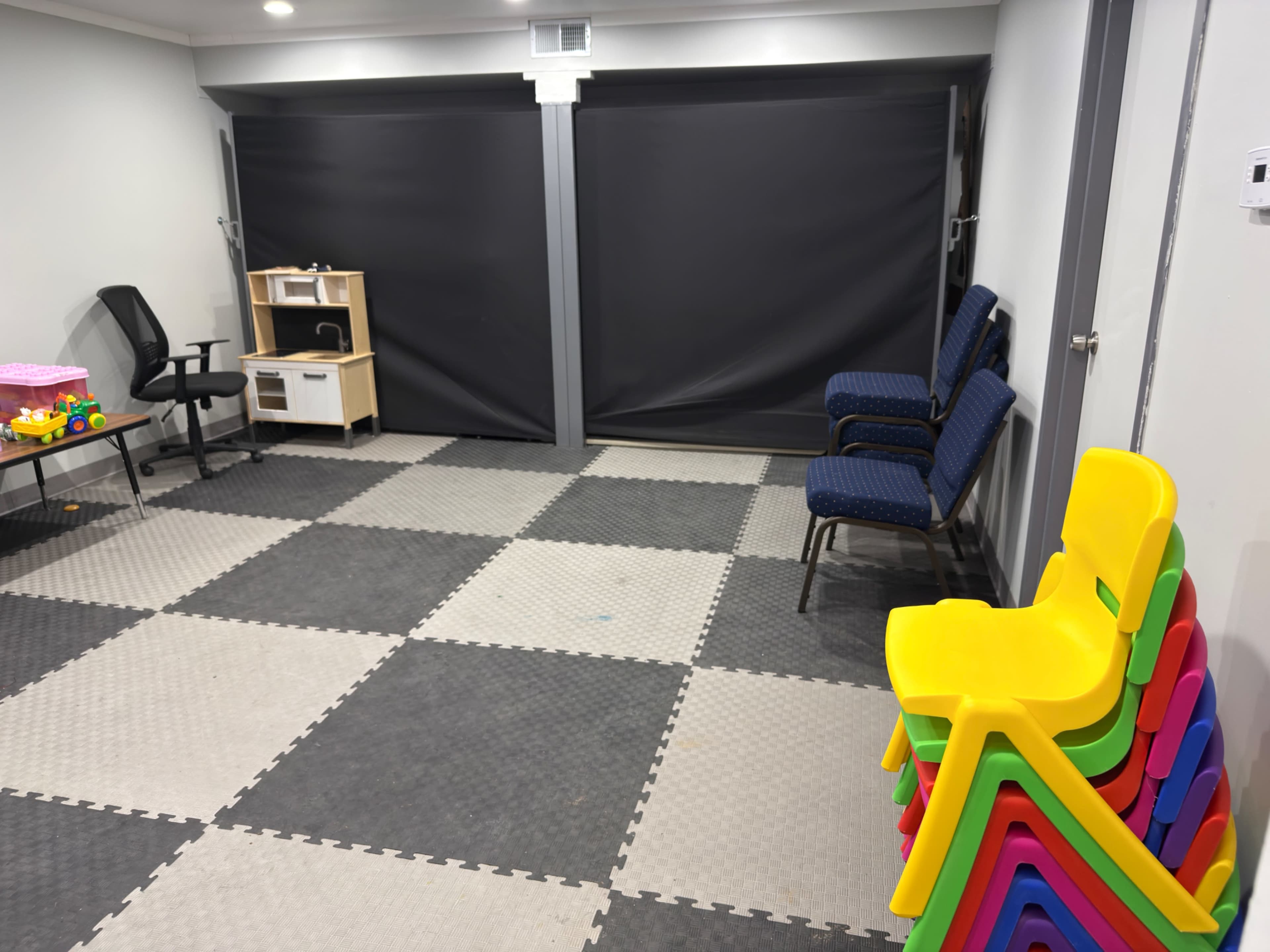 The image shows a playroom with gray and black tile flooring, featuring a chair, a small shelving unit, and a stack of brightly colored plastic chairs.