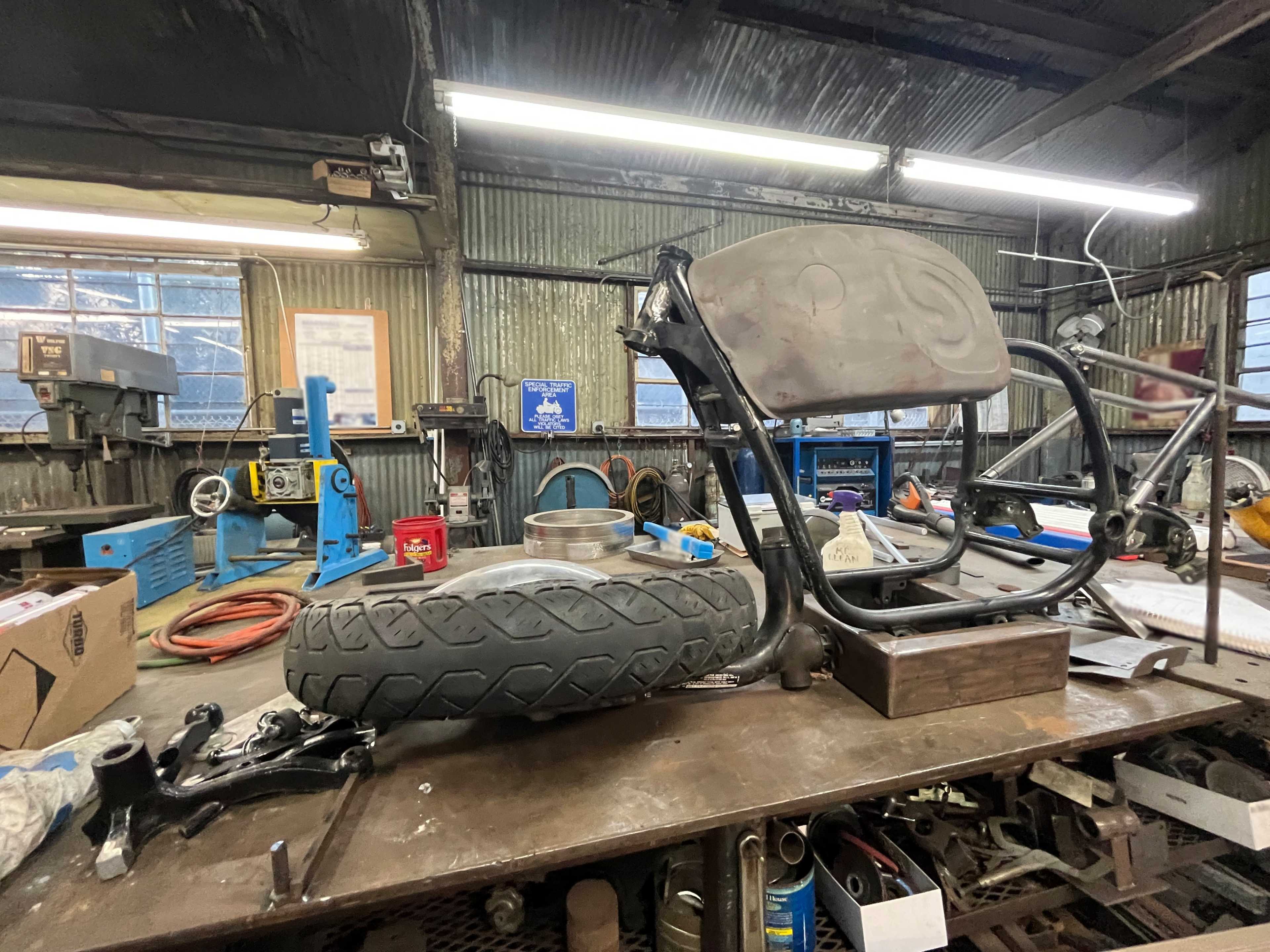 A motorcycle frame with a rear tire is placed on a workbench inside a metal workshop.