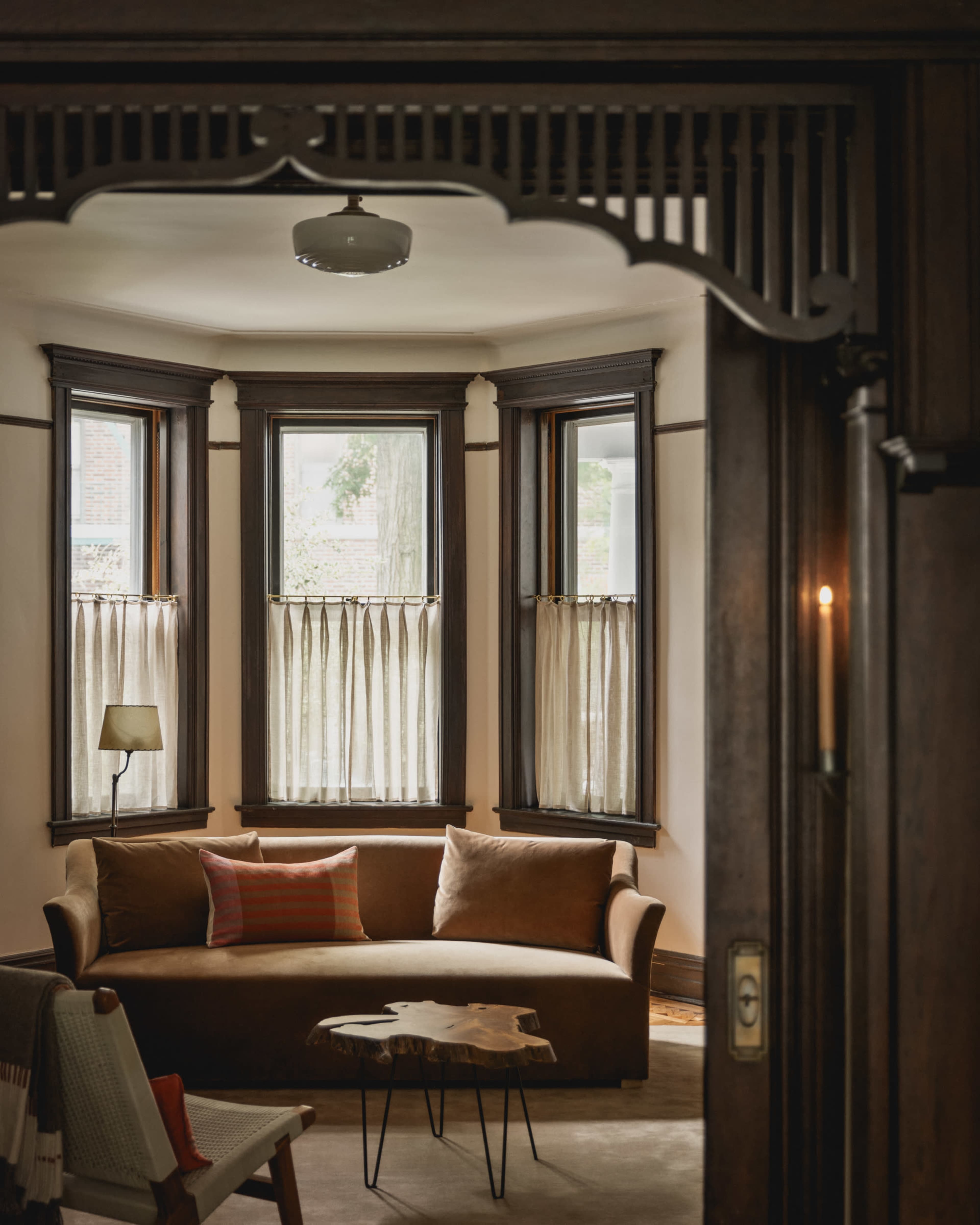 A cozy living room features a brown sofa, a small wooden coffee table, and large windows with sheer curtains.