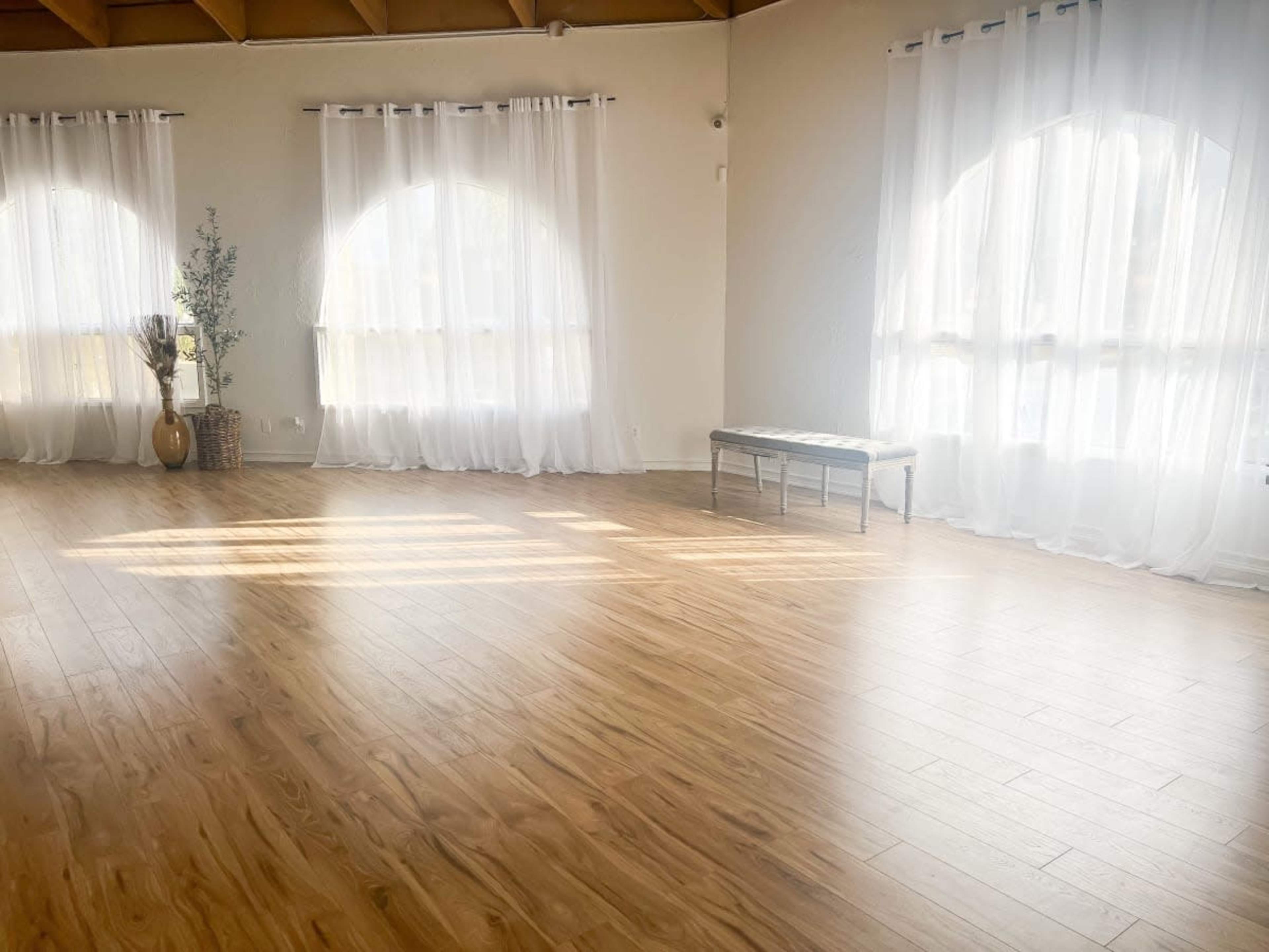 A spacious room with wooden flooring, large windows covered by sheer curtains, and a bench positioned in the center.