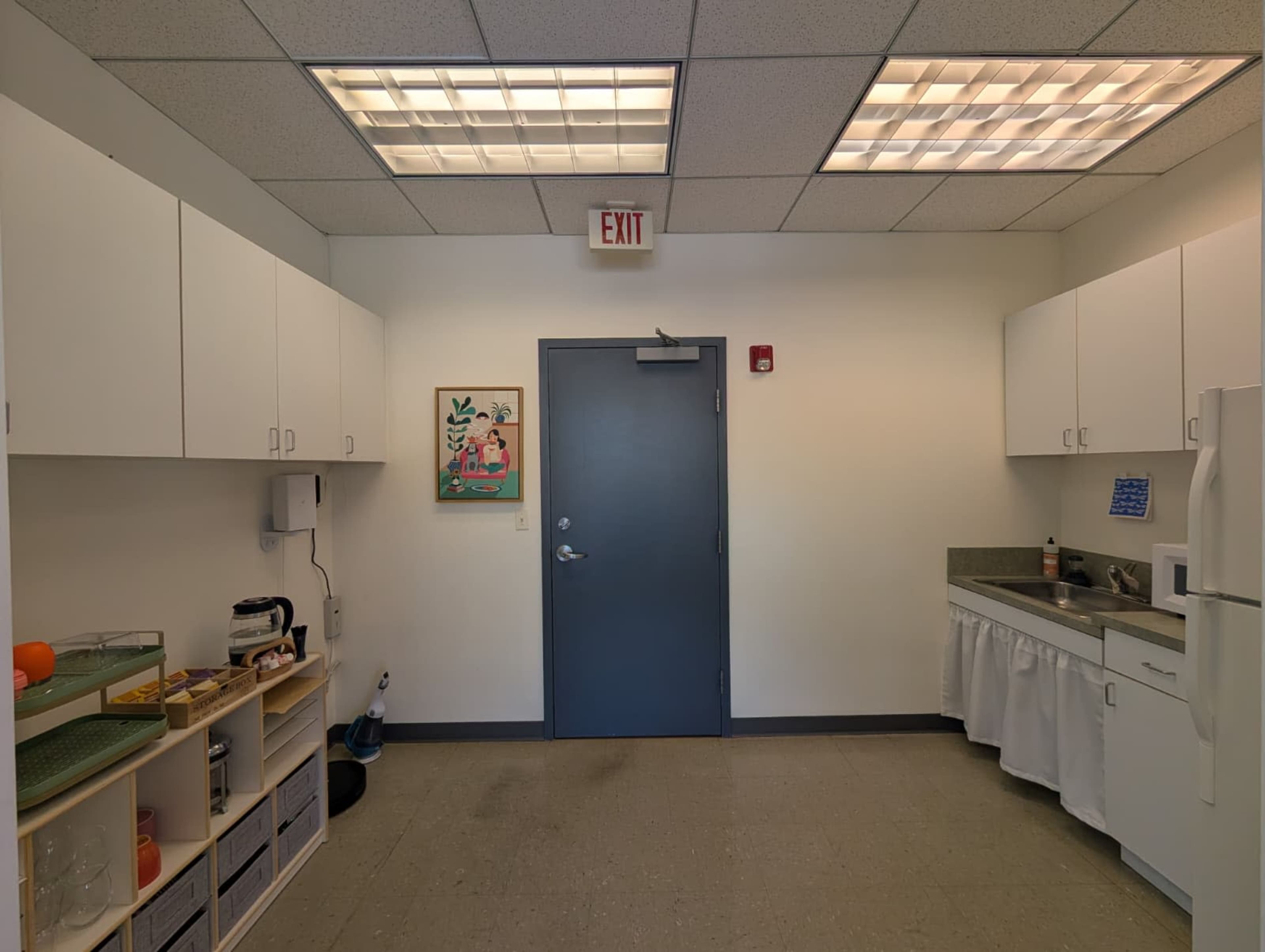 The image shows a simple kitchen area with cabinets, a countertop, a refrigerator, and a door leading to another space, illuminated by ceiling lights.