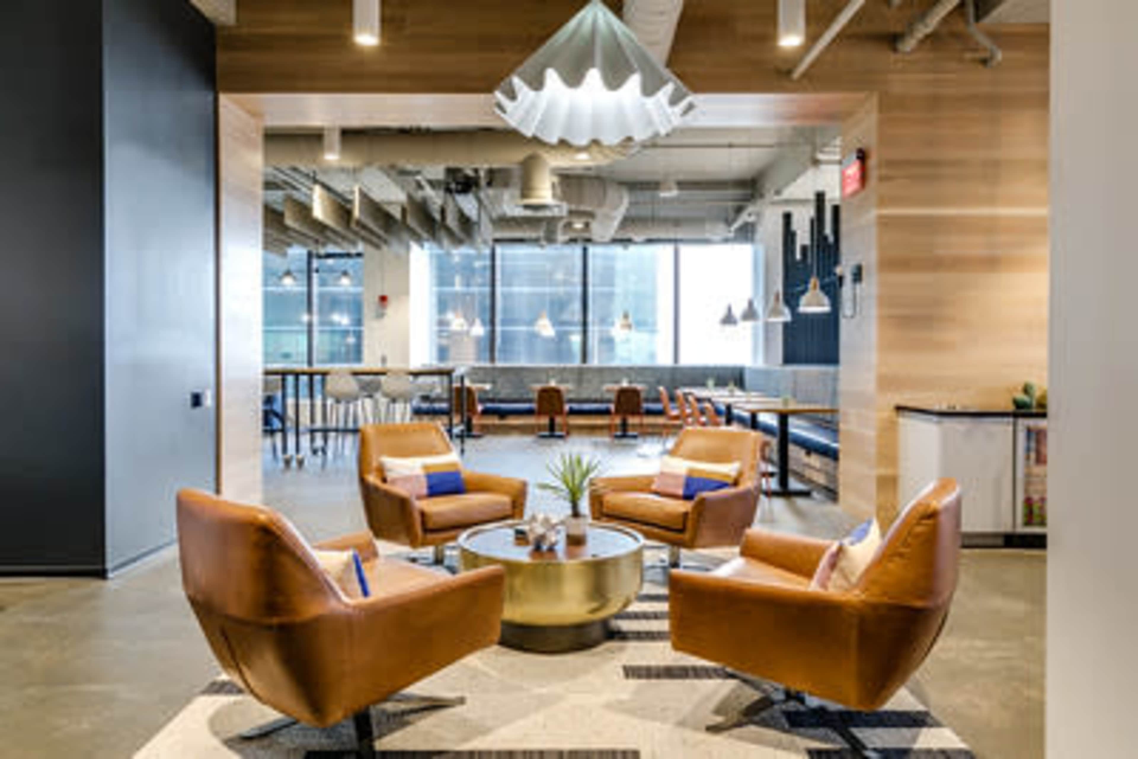 The image shows a modern lounge area featuring four brown leather chairs arranged around a circular coffee table, with large windows and contemporary lighting fixtures in the background.
