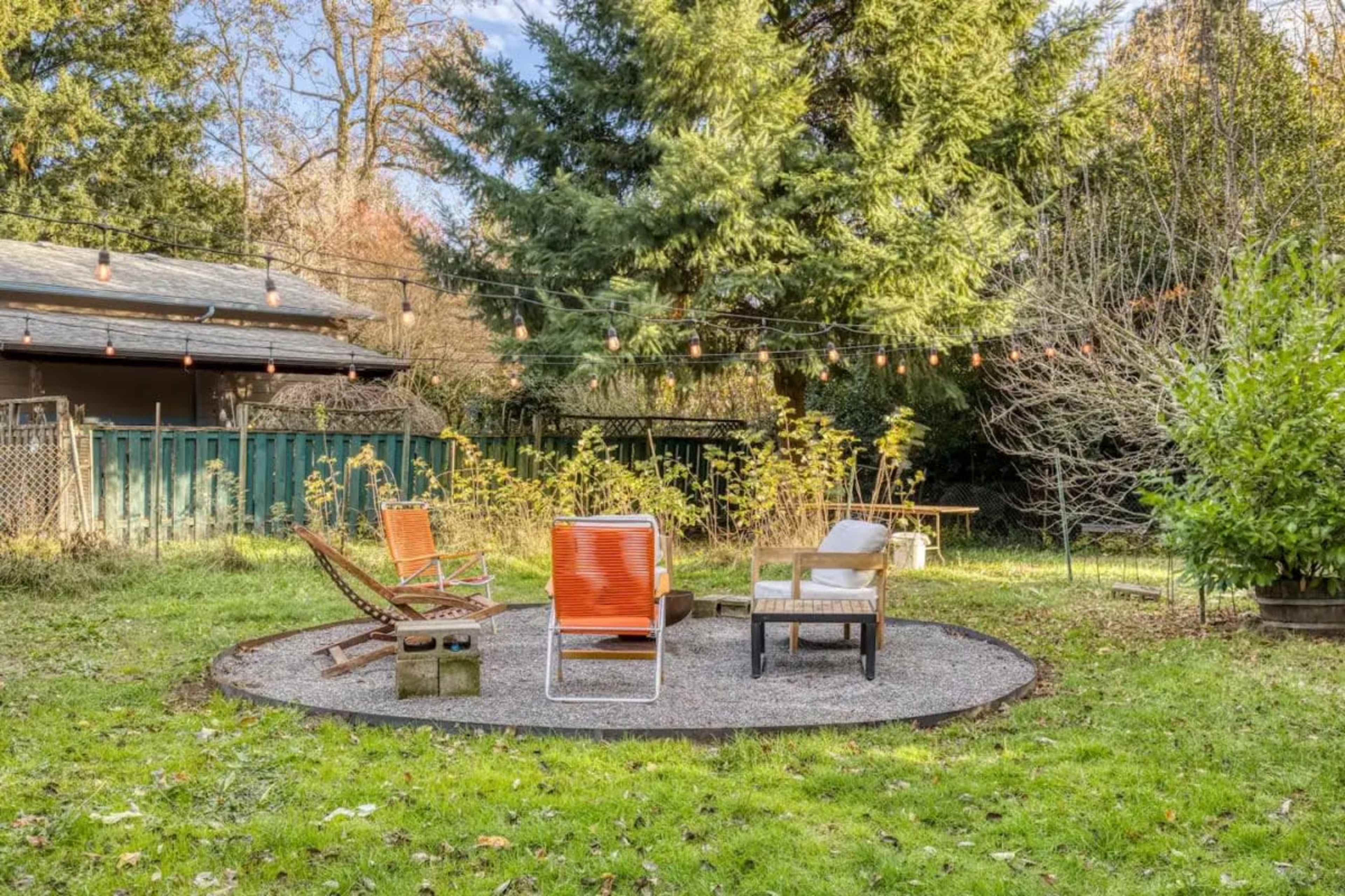 A circular gravel sitting area features orange chairs and a small table, surrounded by greenery and string lights in a backyard.
