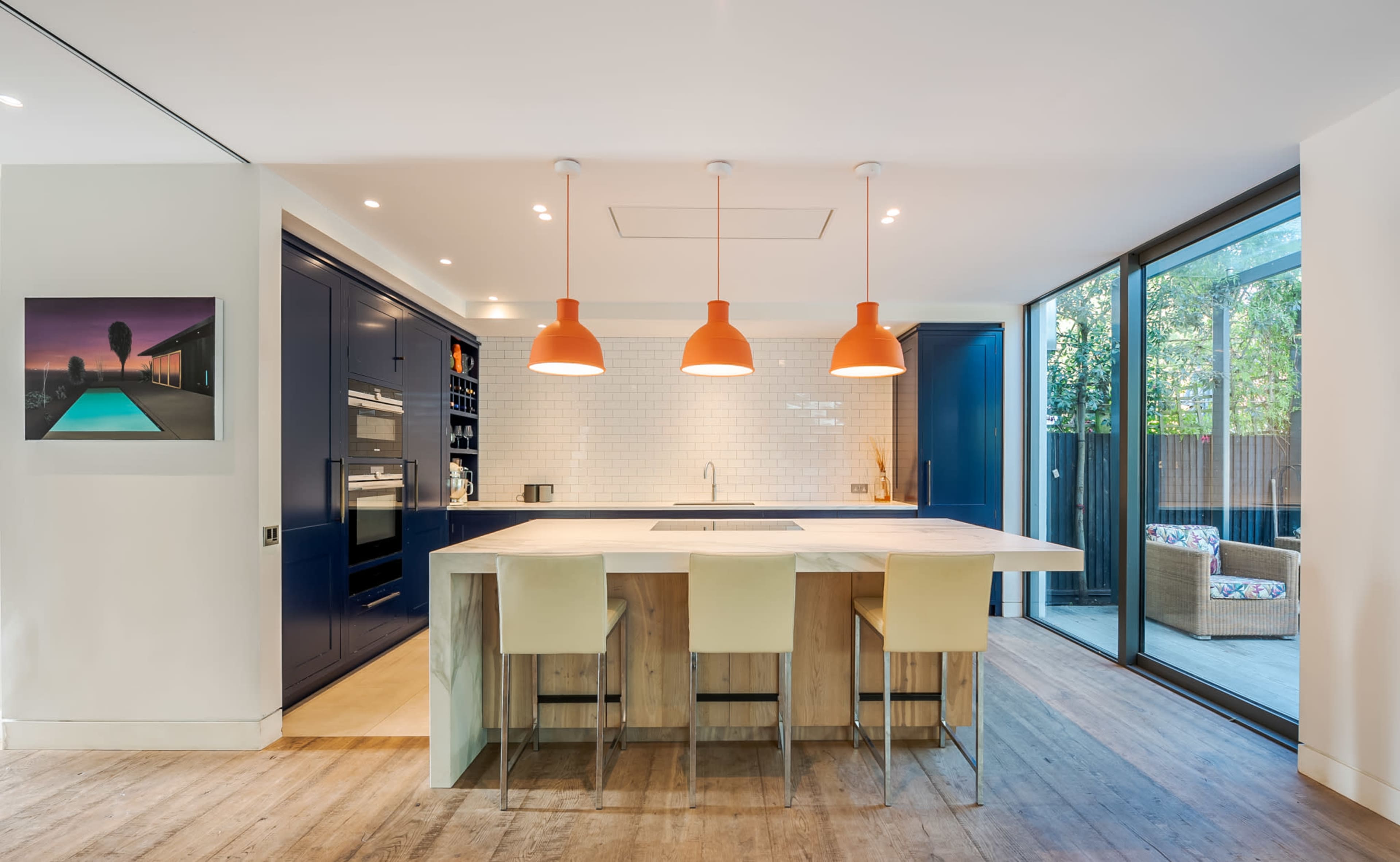 A modern kitchen features a central island with bar stools, orange pendant lights, and floor-to-ceiling windows.