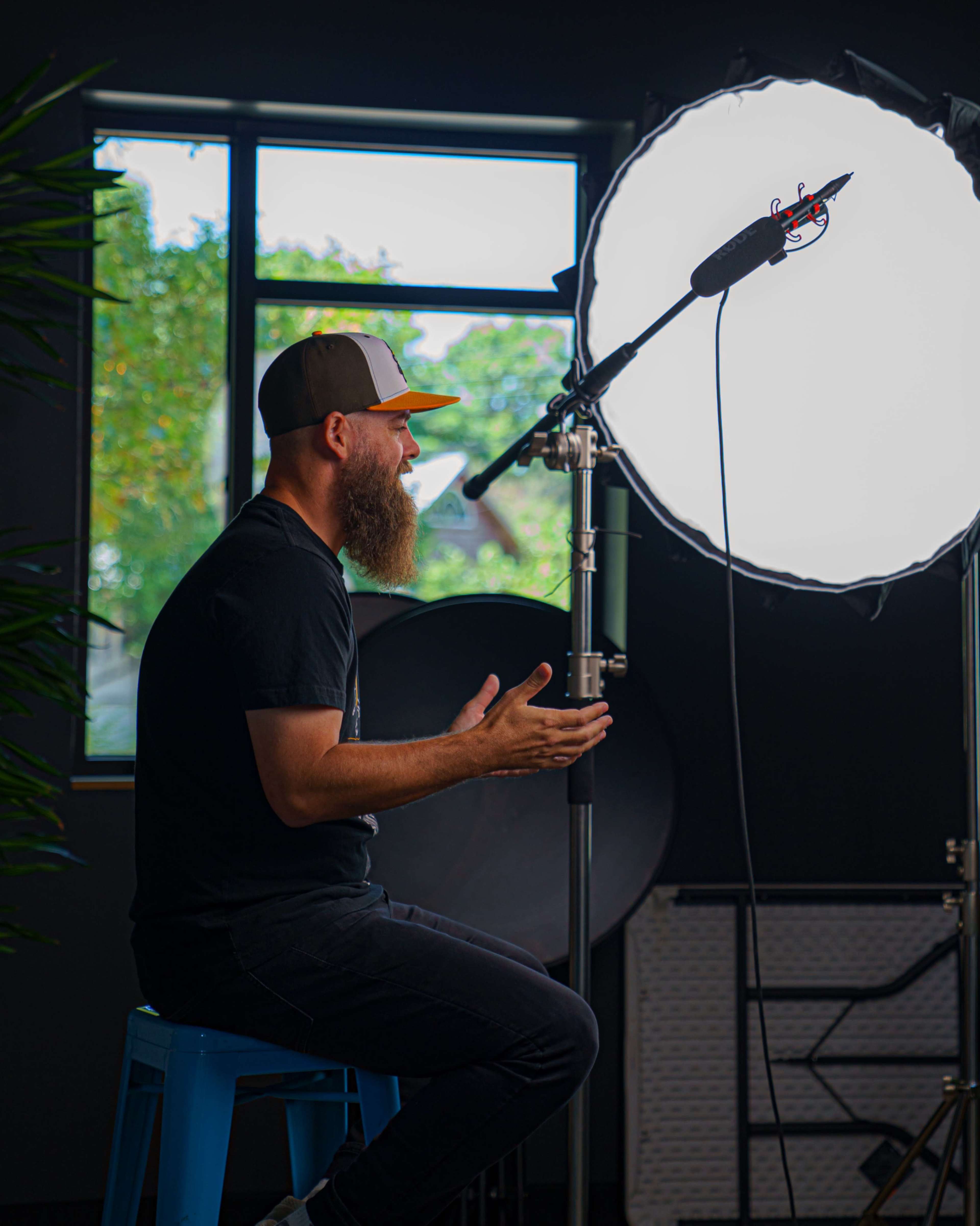 A man with a beard sits on a stool in front of a large light source, gesturing as he speaks.