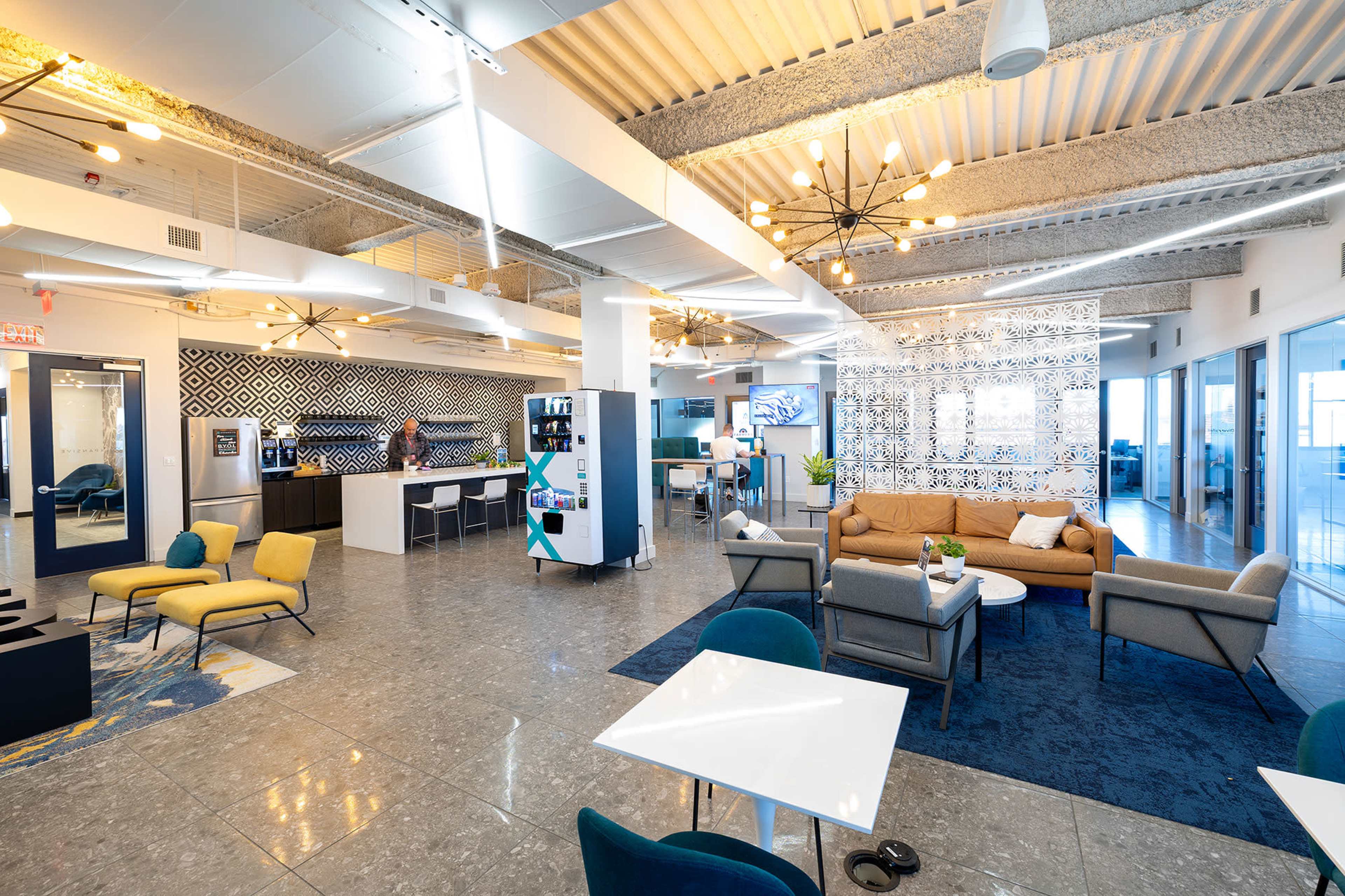 The image shows a bright, modern coworking space with various seating areas, a kitchen, and decorative wall patterns.