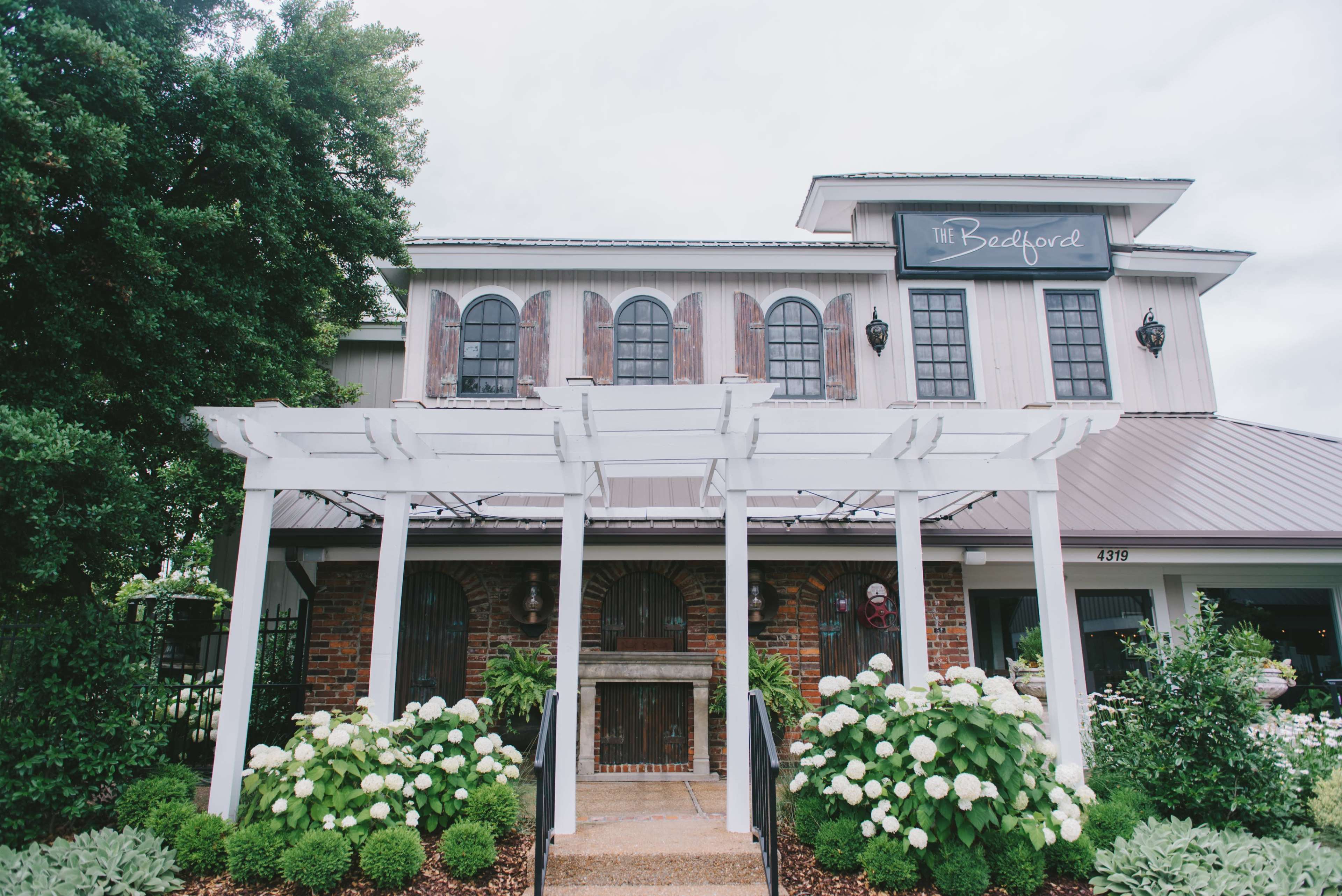 A restaurant building with a white pergola entrance, surrounded by green shrubs and flowers, under a cloudy sky.