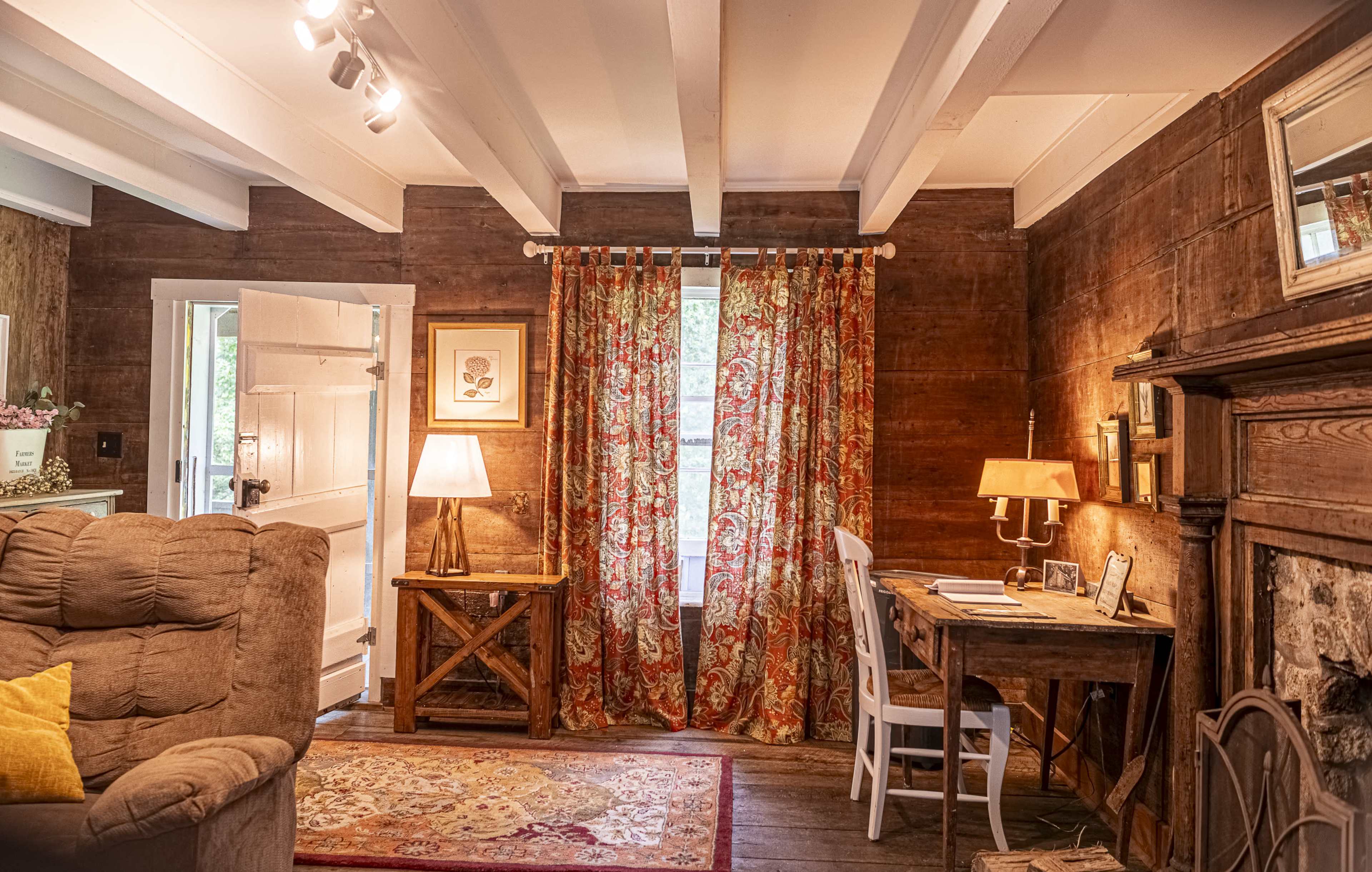 A cozy room with wooden walls, a sofa, a desk, a lamp, and floral curtains framing a window.