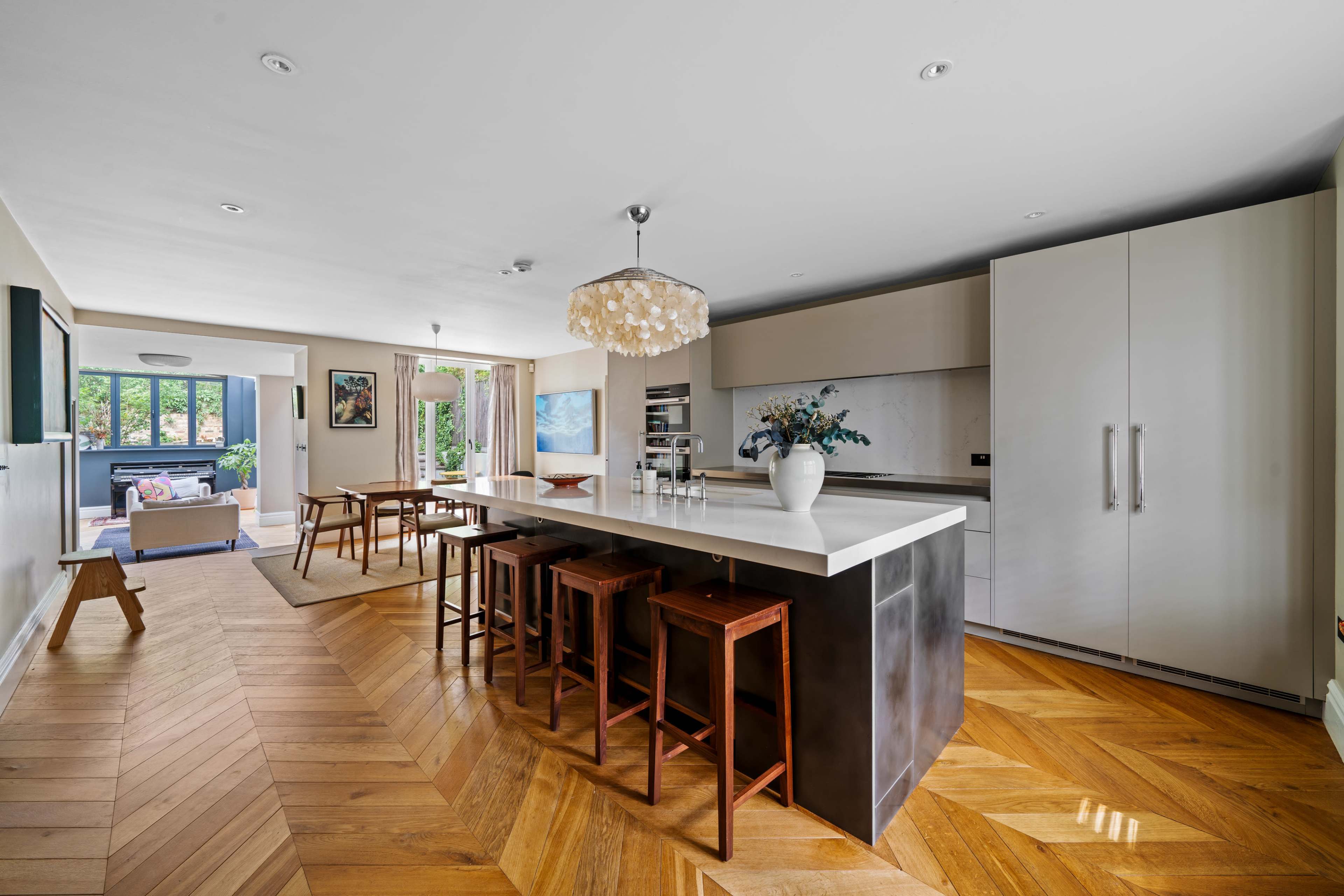 A modern kitchen features a large island with high stools, sleek cabinetry, and a view into a living area with natural light.