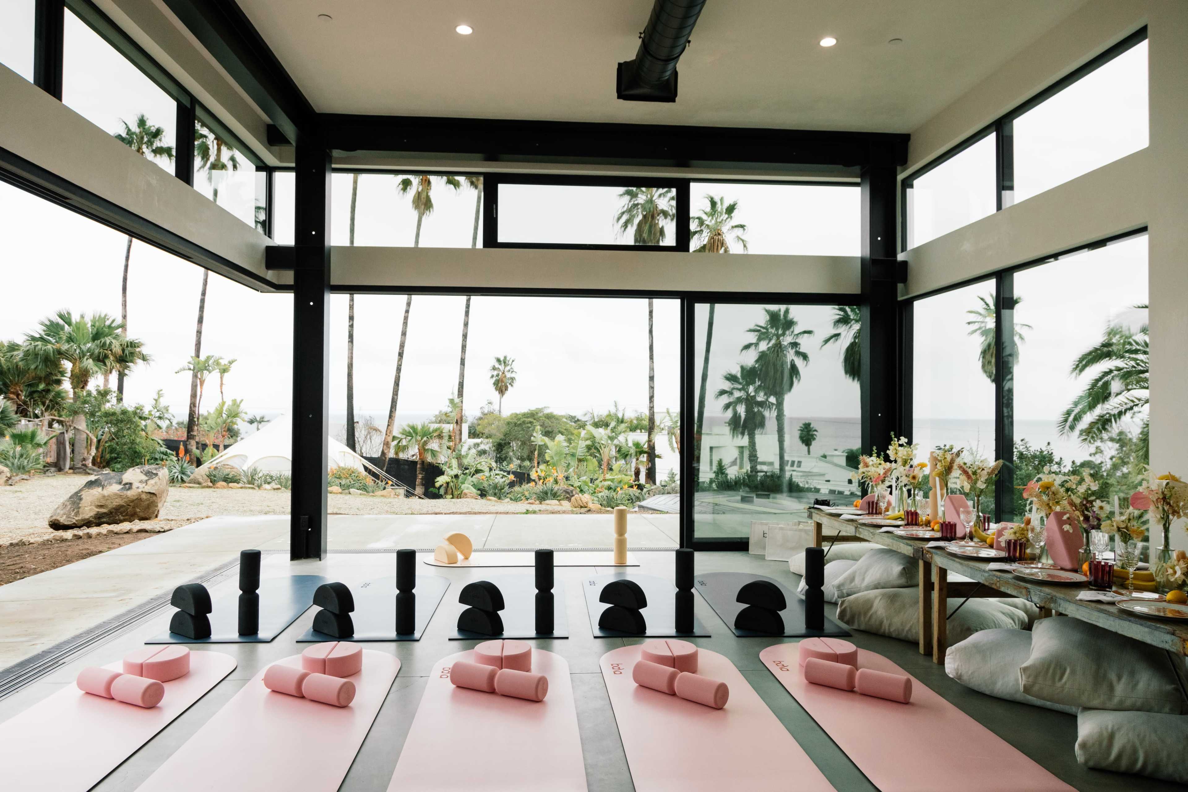 The image shows a spacious yoga studio with pink mats arranged in rows and large windows overlooking a tropical landscape with palm trees.