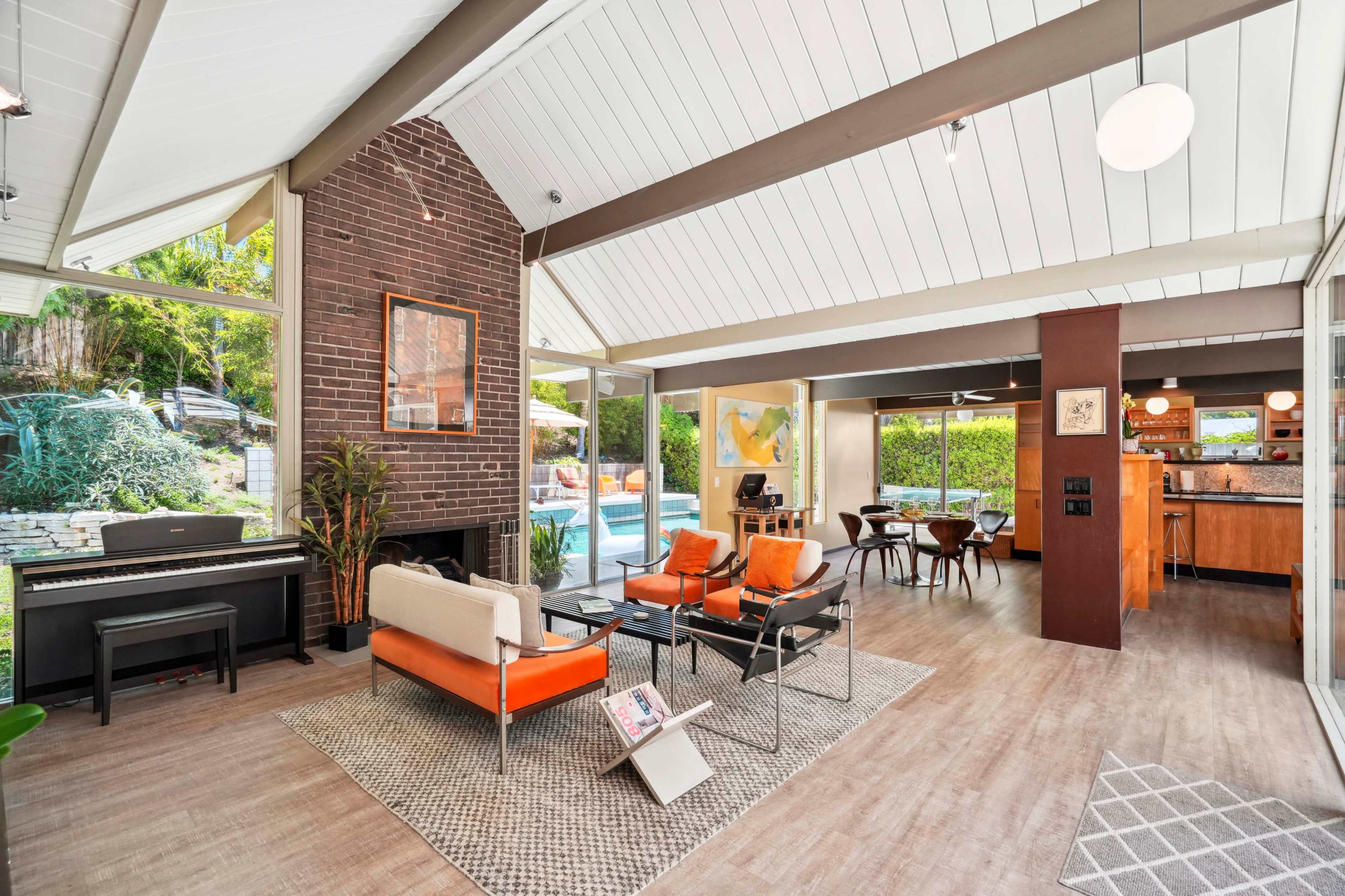 A spacious living area with large windows, featuring orange chairs, a piano, and an adjacent kitchen visible in the background.