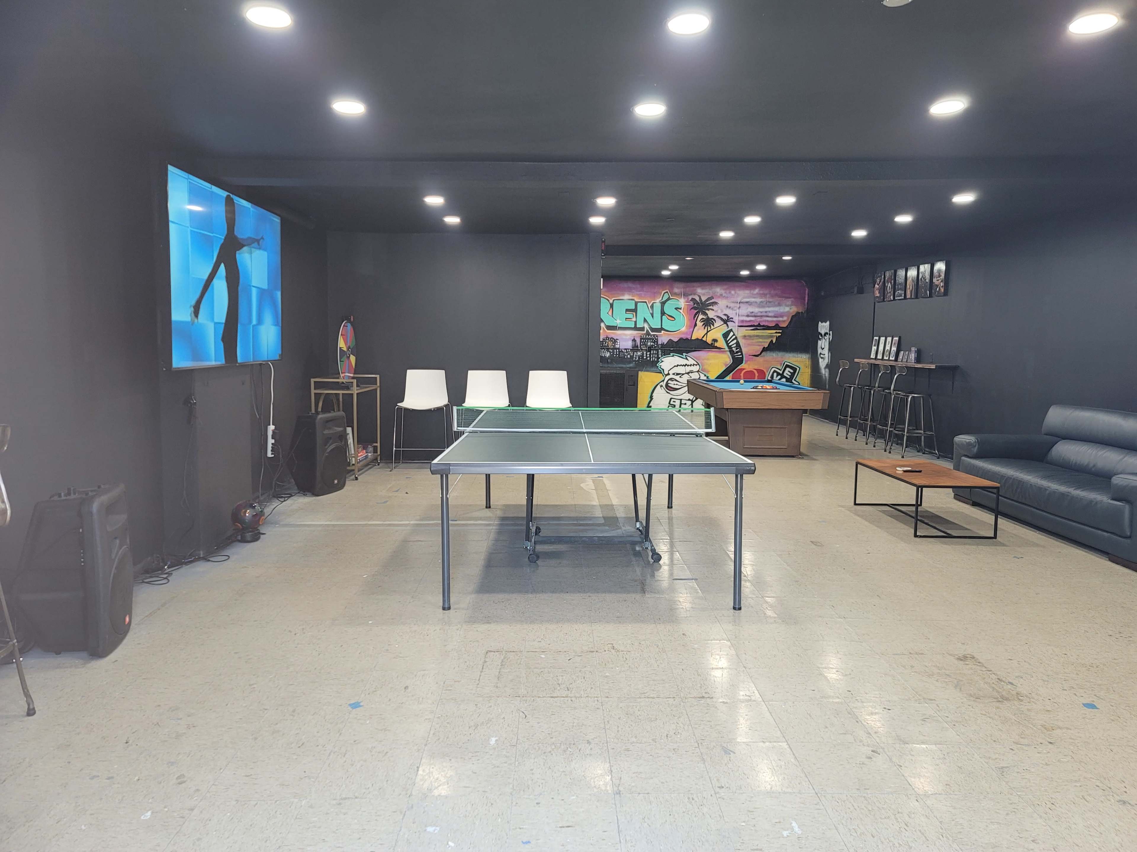 The image shows a room featuring a ping pong table, white chairs, a couch, a television screen, and a colorful mural on the wall.