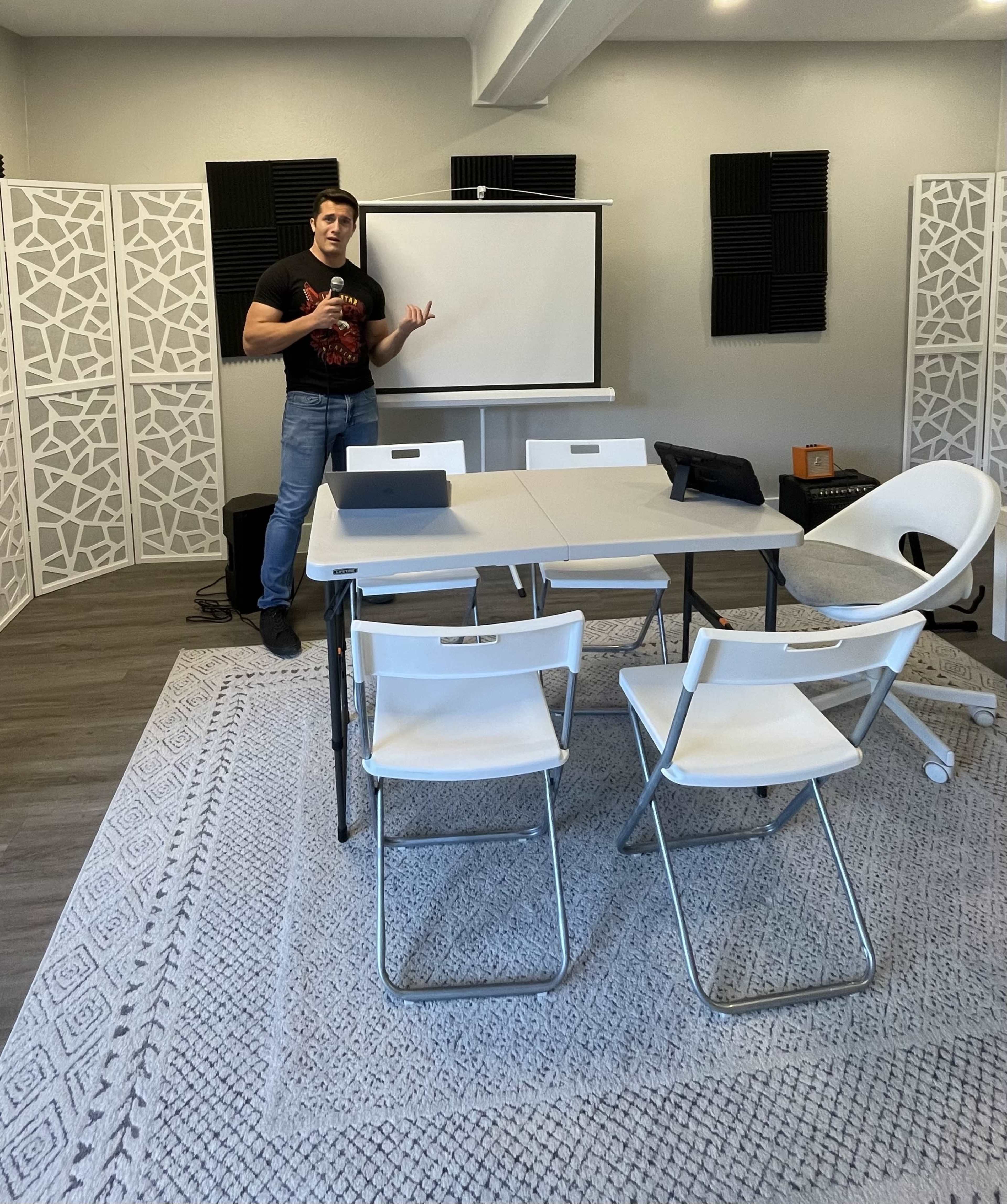 A person stands beside a projection screen in a modern meeting room with a table and chairs arranged for a presentation.