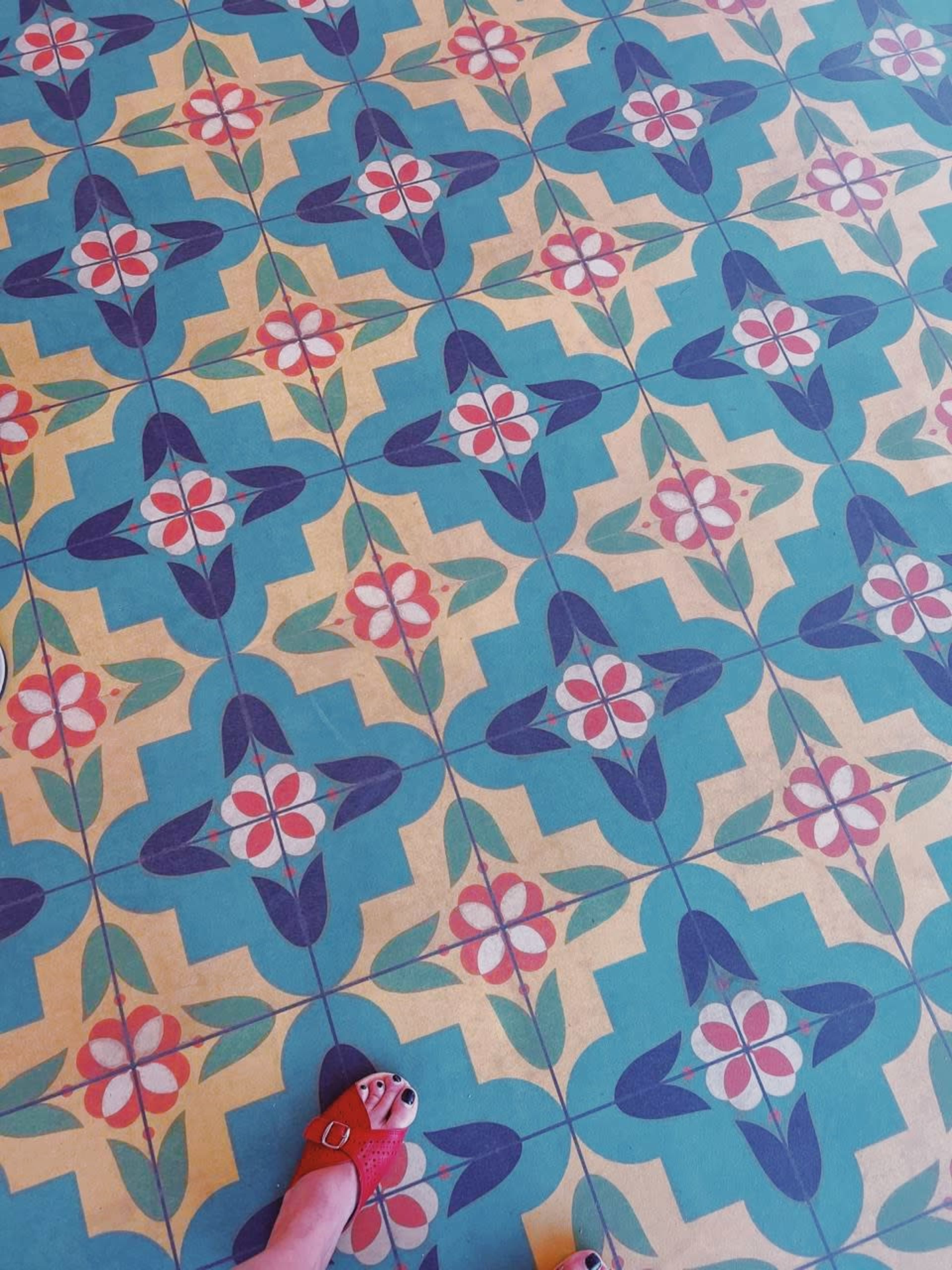 A patterned floor covering featuring colorful floral designs in a geometric layout, with a portion of a person's foot in red sandals visible at the bottom.