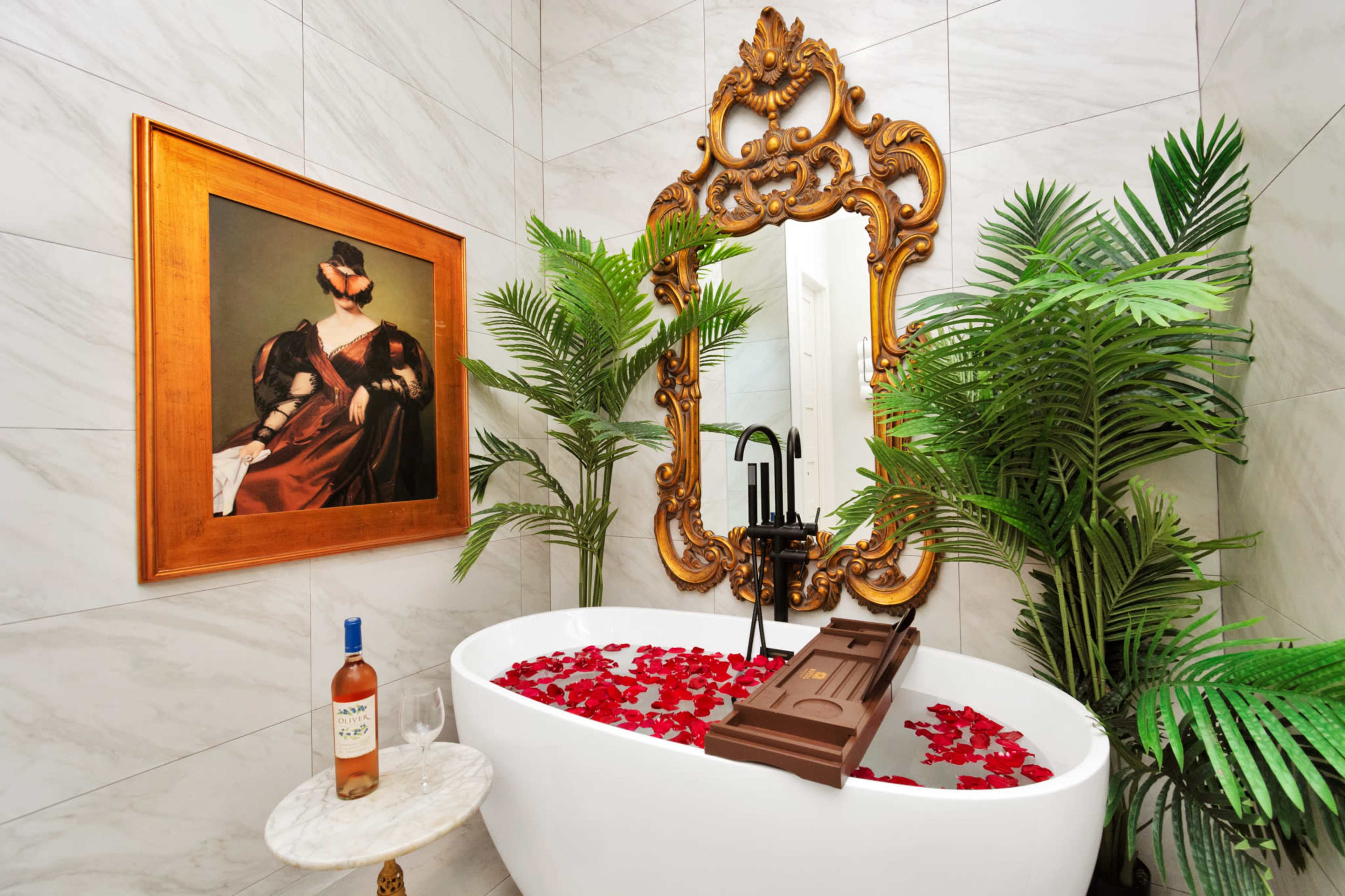 A bathtub filled with rose petals is accompanied by a framed painting and greenery in a stylish bathroom setting.