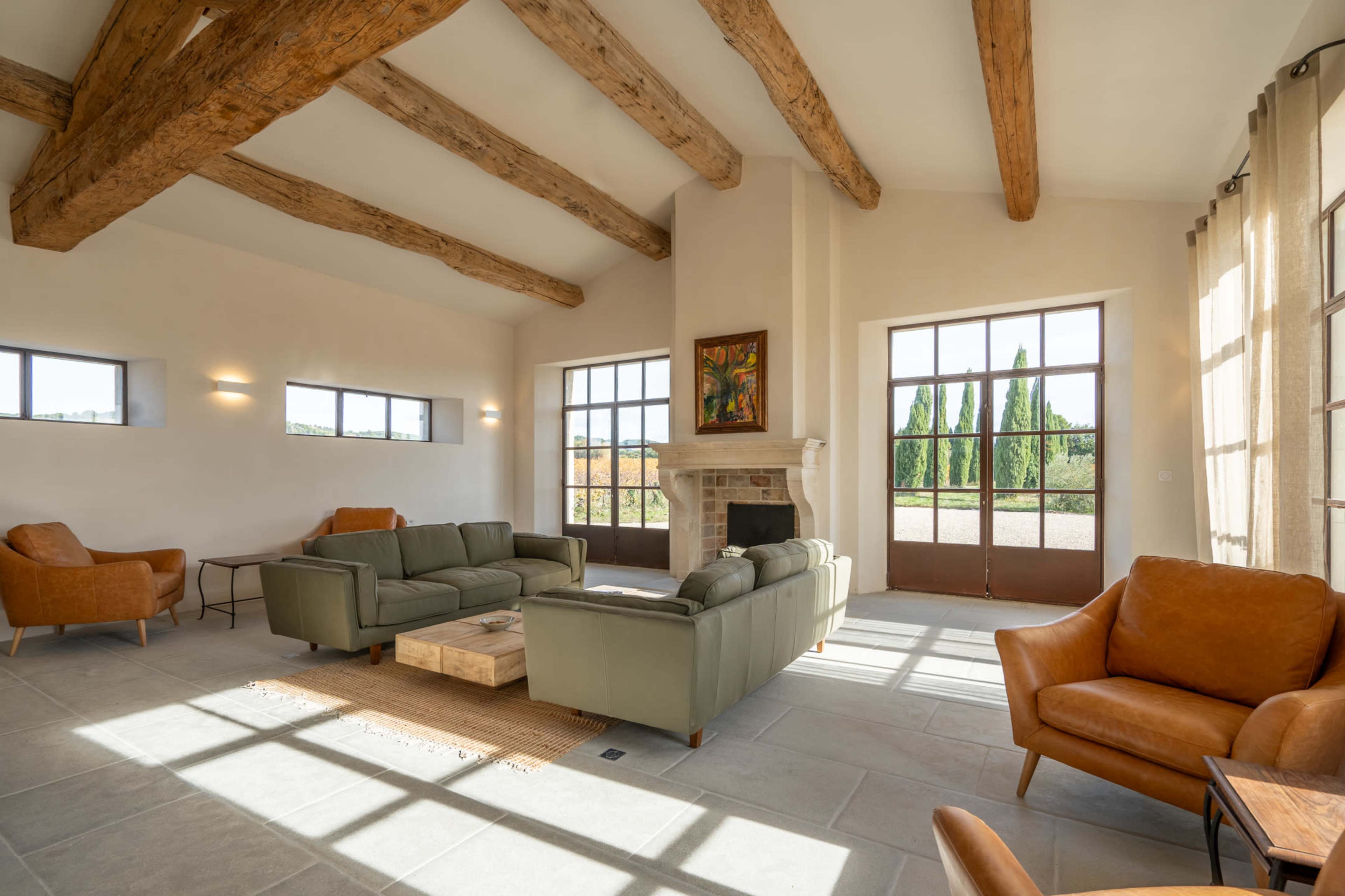 The image shows a spacious living room with wooden beams, large windows, a fireplace, and a mix of green and brown furniture.