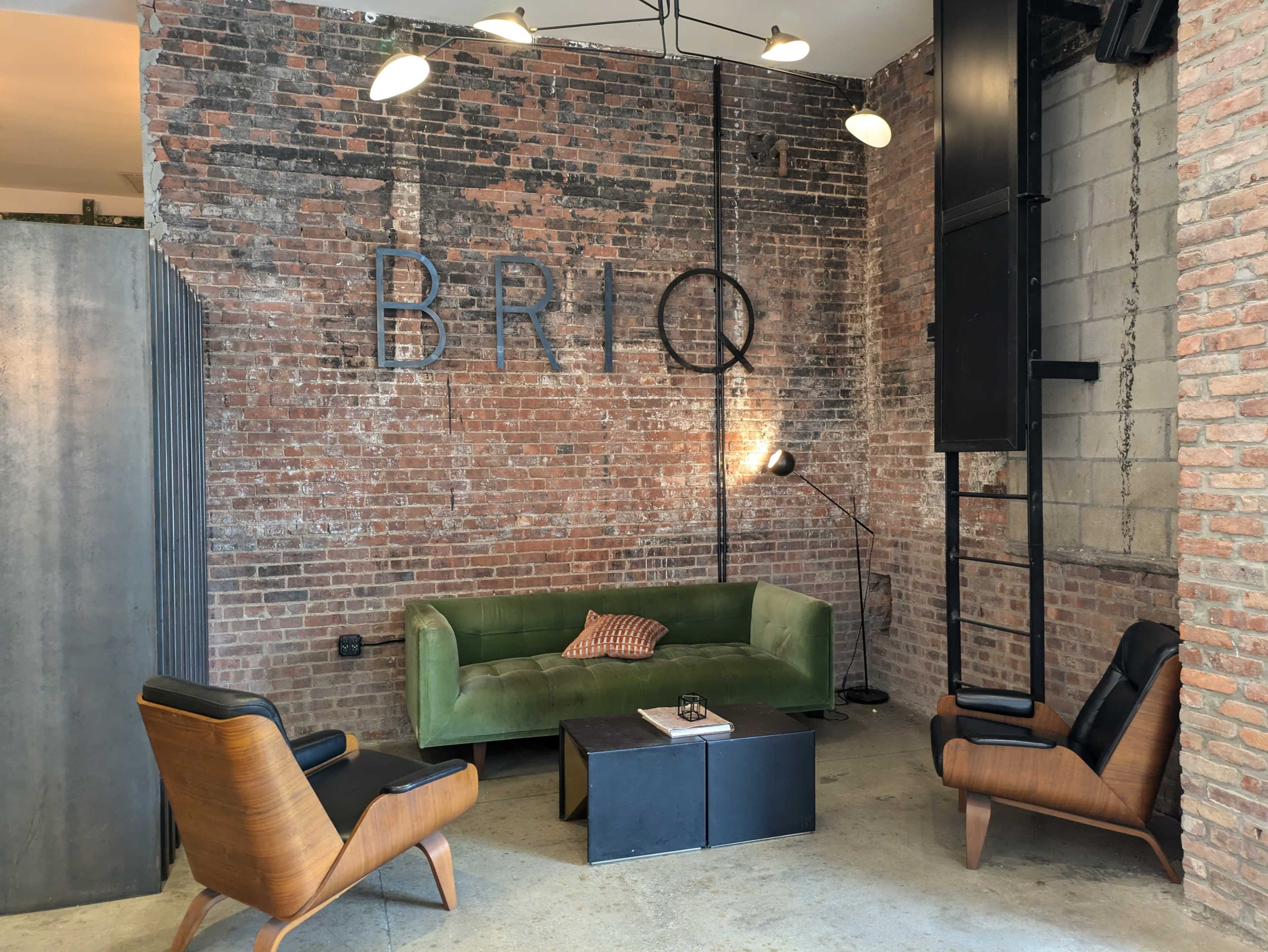 The image shows a modern lounge area featuring a green velvet sofa, two black armchairs, a coffee table, and a brick wall with the word "BRIQ" displayed on it.