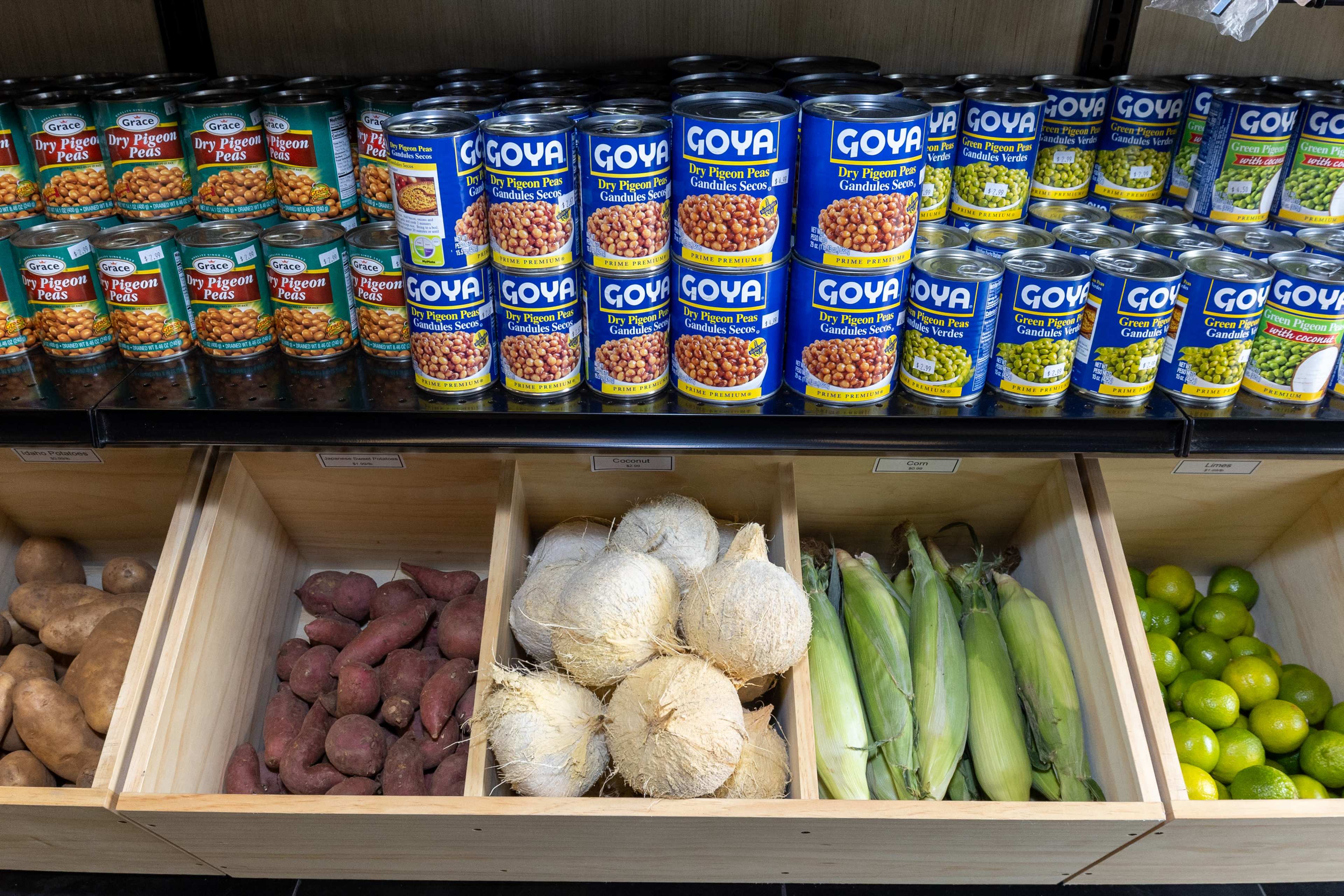 A grocery store display features shelves with canned goods labeled Goya and wooden bins containing sweet potatoes, root vegetables, corn, and limes.