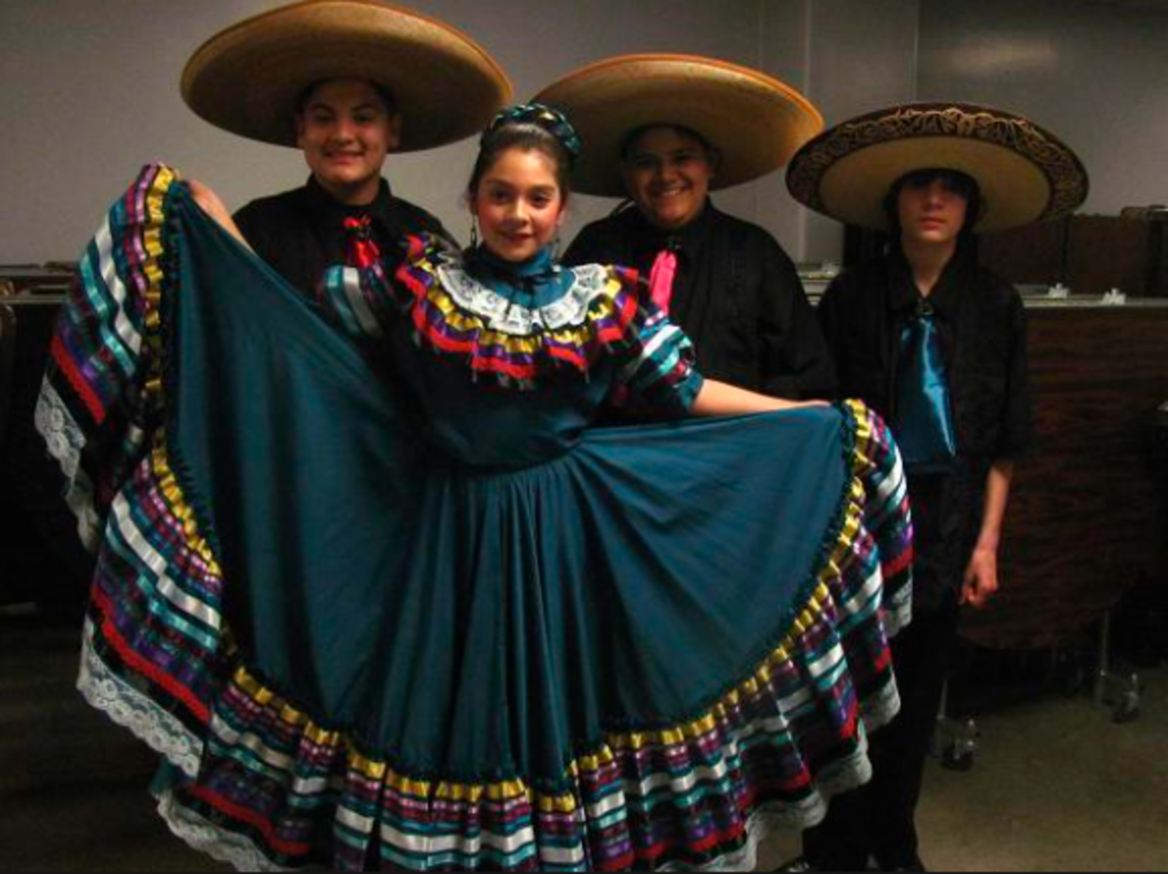 Four individuals wearing traditional Mexican attire, including large sombreros and colorful dresses, pose together in a indoor setting.