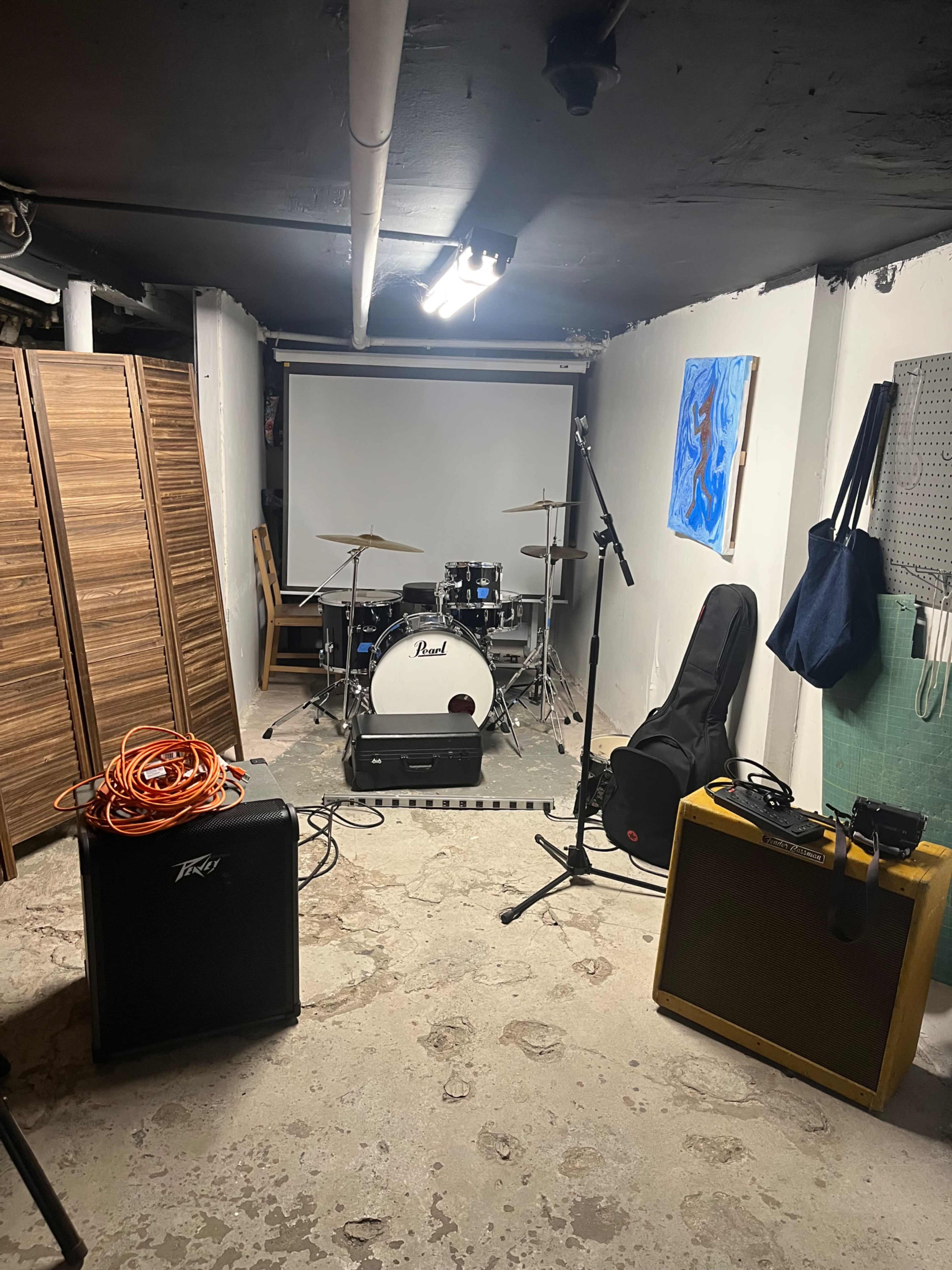 The image shows a basement music rehearsal space with a drum set, an amplifier, a guitar case, and a projection screen on the wall.